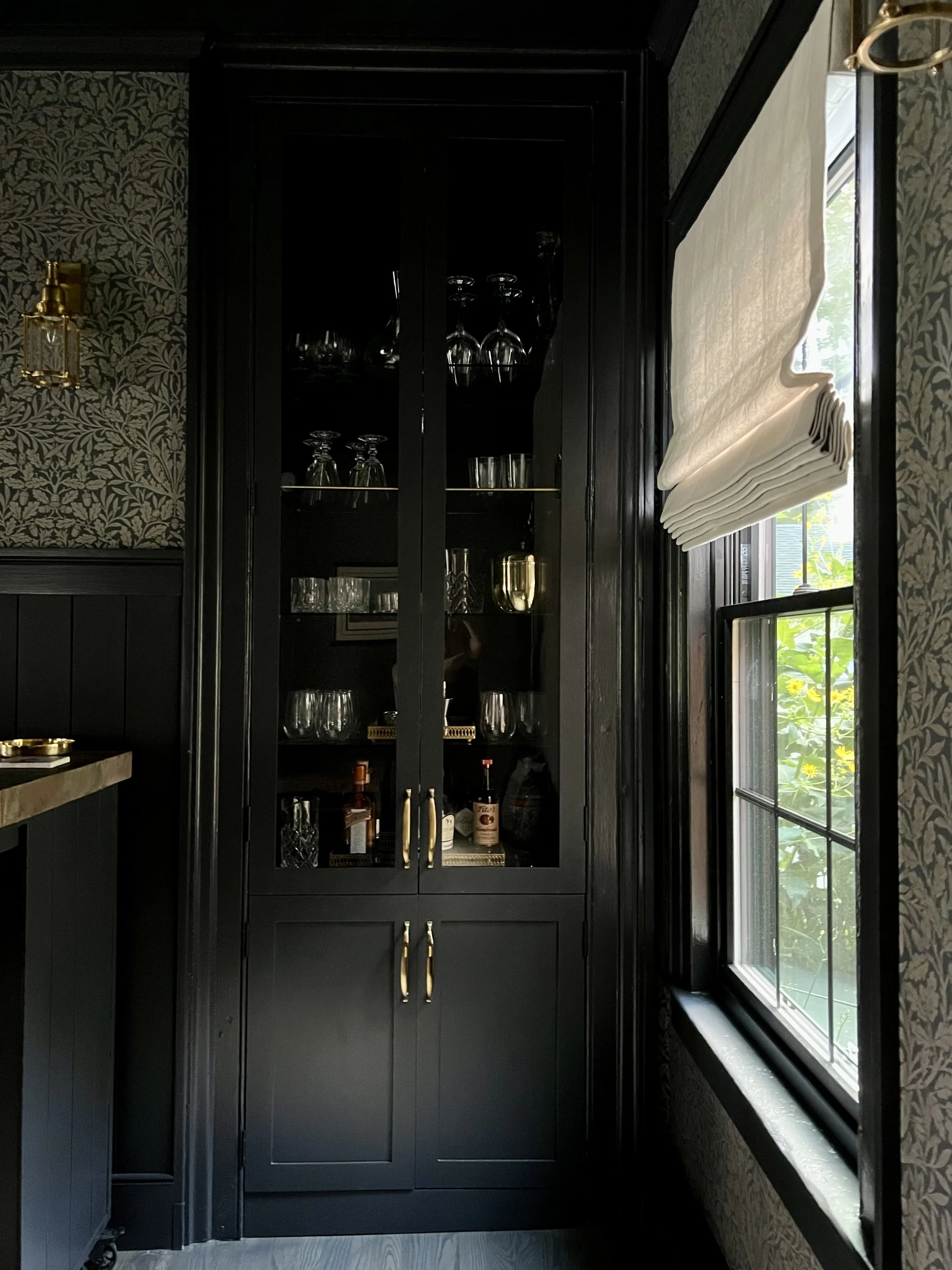 A black kitchen cabinet with glass doors displaying glassware, bottles, and bar accessories, next to a window with a partially rolled white blind, in a room with dark patterned wallpaper.
