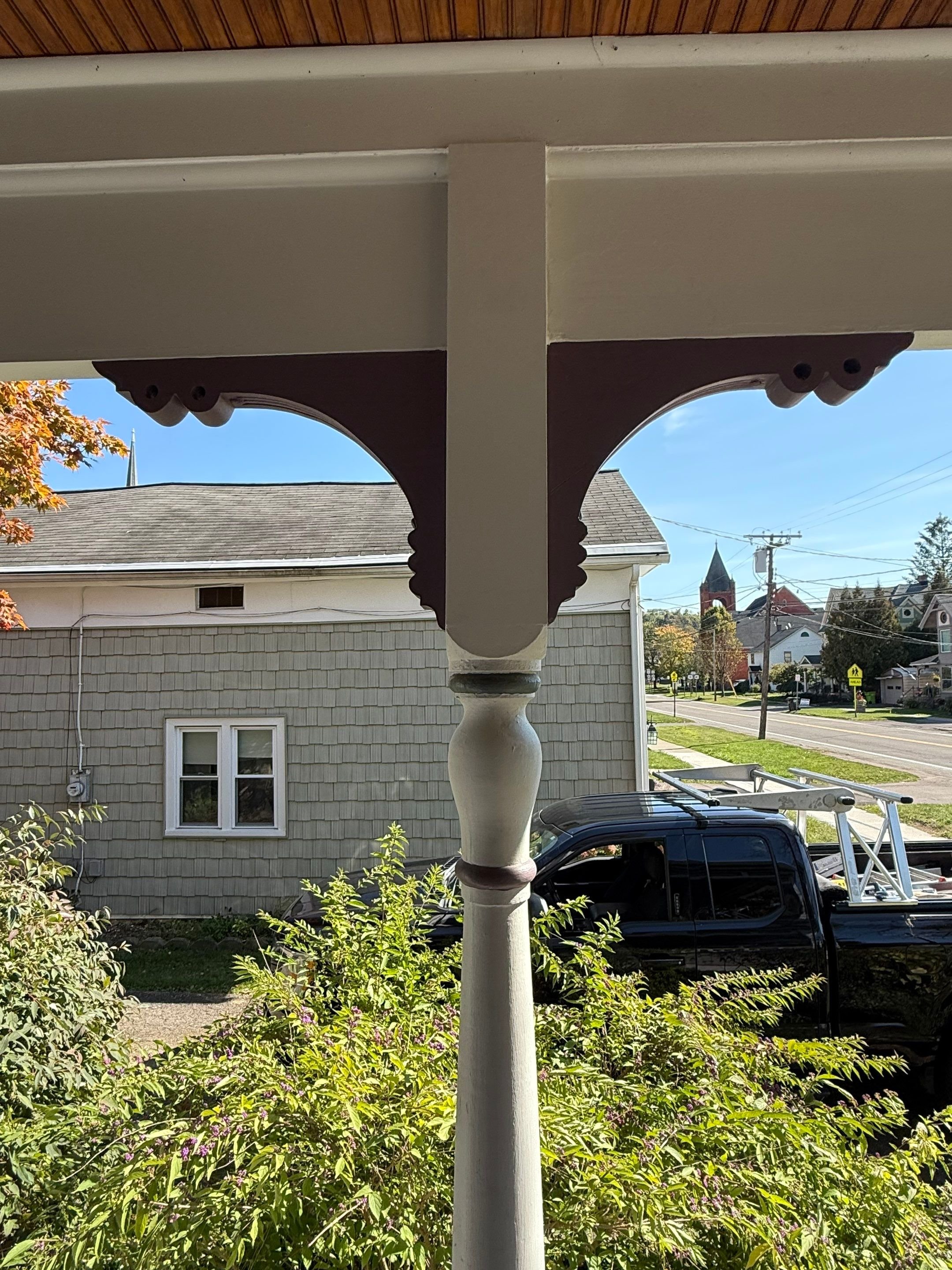 View from a porch showing an ornate wooden porch post, beige house with shingle siding, a black pickup truck with a ladder, and a street with a church steeple and trees in the background.