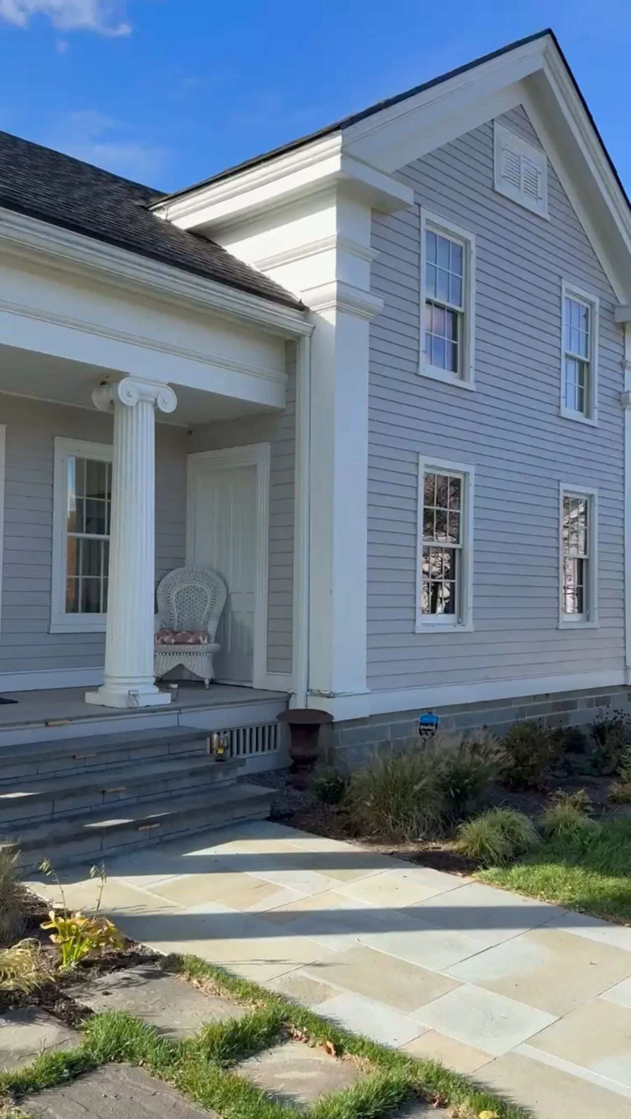 Front porch of a two-story house with a classical column, light gray shingles, and a stone walkway leading to stairs.