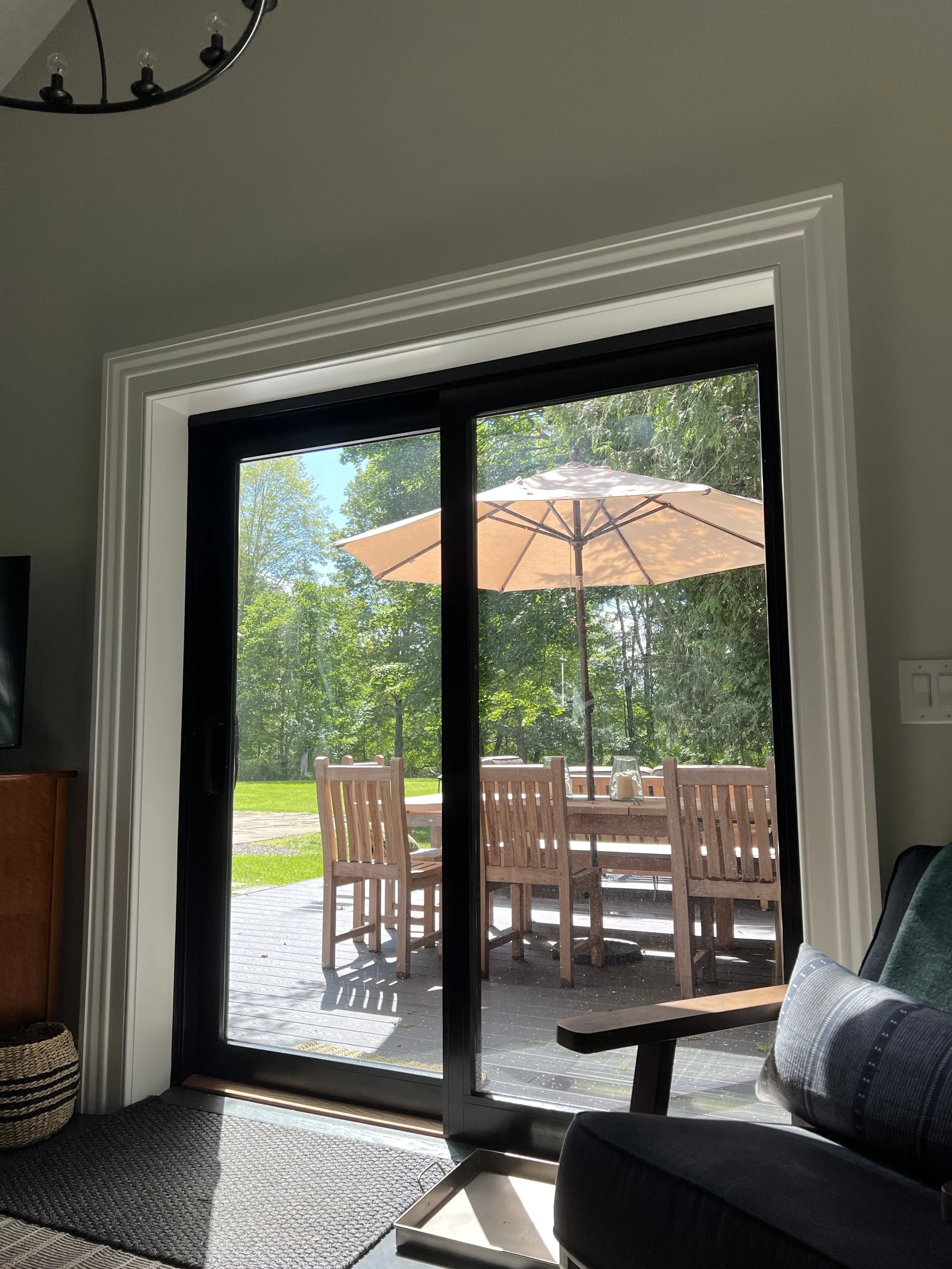 View through sliding glass door showing outdoor wooden patio furniture and a large beige patio umbrella in a green, wooded backyard.