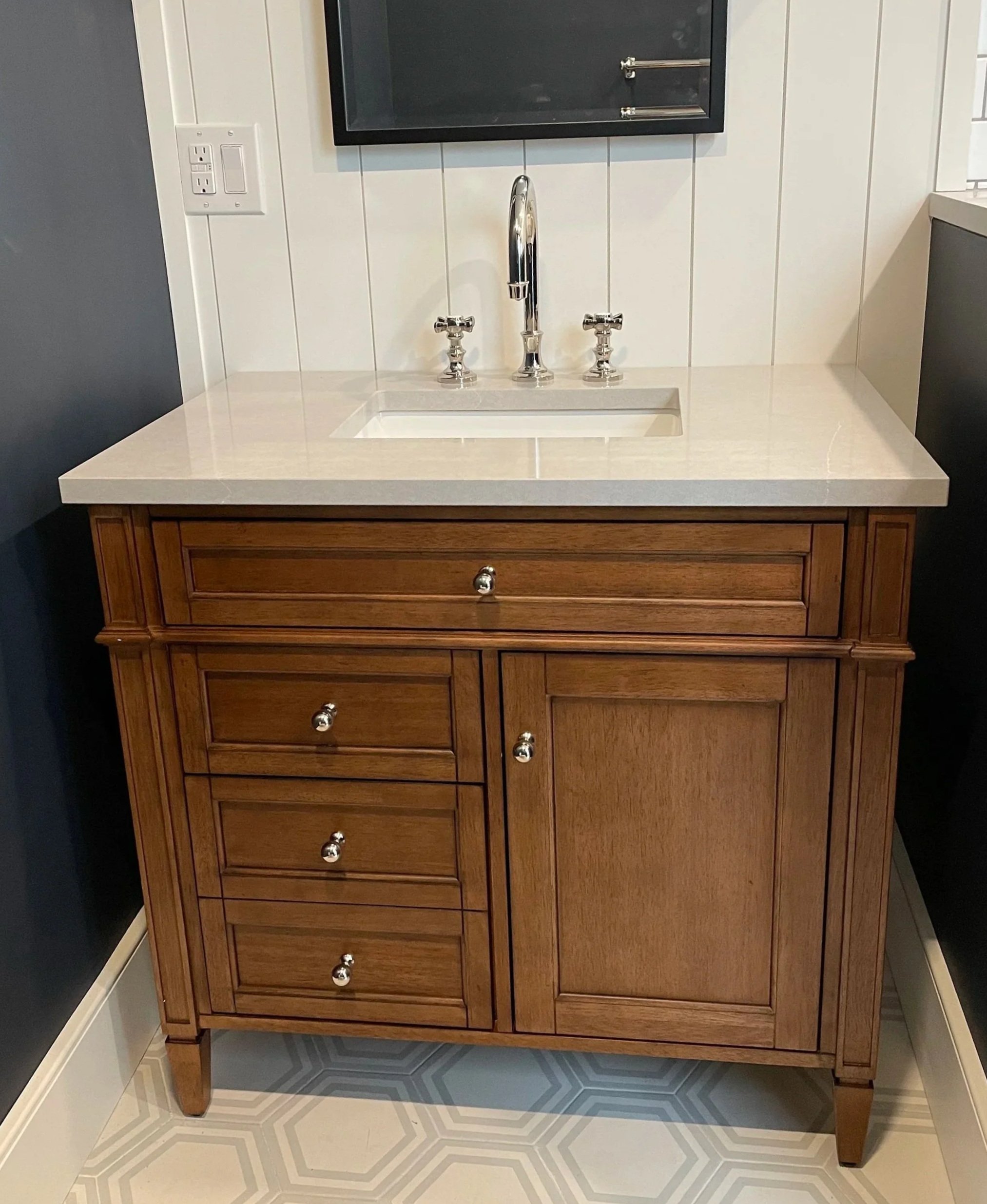 Bathroom wooden vanity with a white countertop, built-in sink, and a chrome faucet, set against white paneled walls with a mirror above.