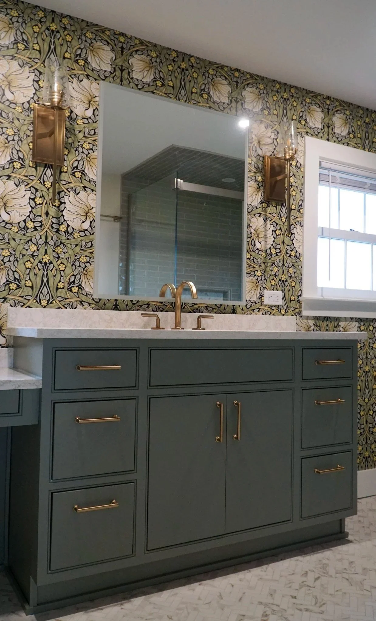 A bathroom countertop with green cabinets and gold handles. Two light fixtures surround a mirror on the wall which reflects a modern shower in the background. Dramatic green wallpaper with white flowers and black accents.