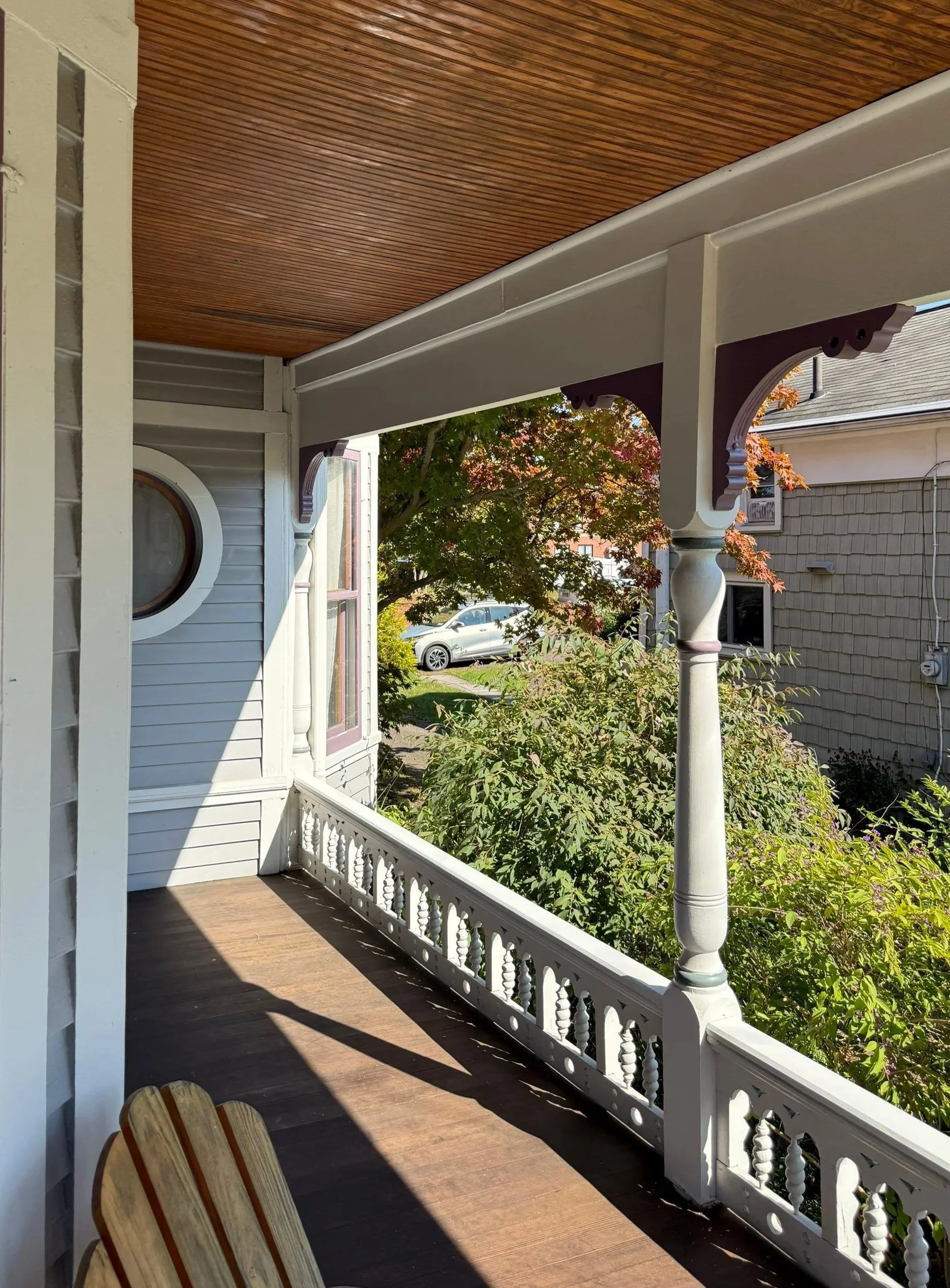 Front porch of a house with wooden flooring, decorative railing, supporting columns, and a view of trees and neighboring houses.