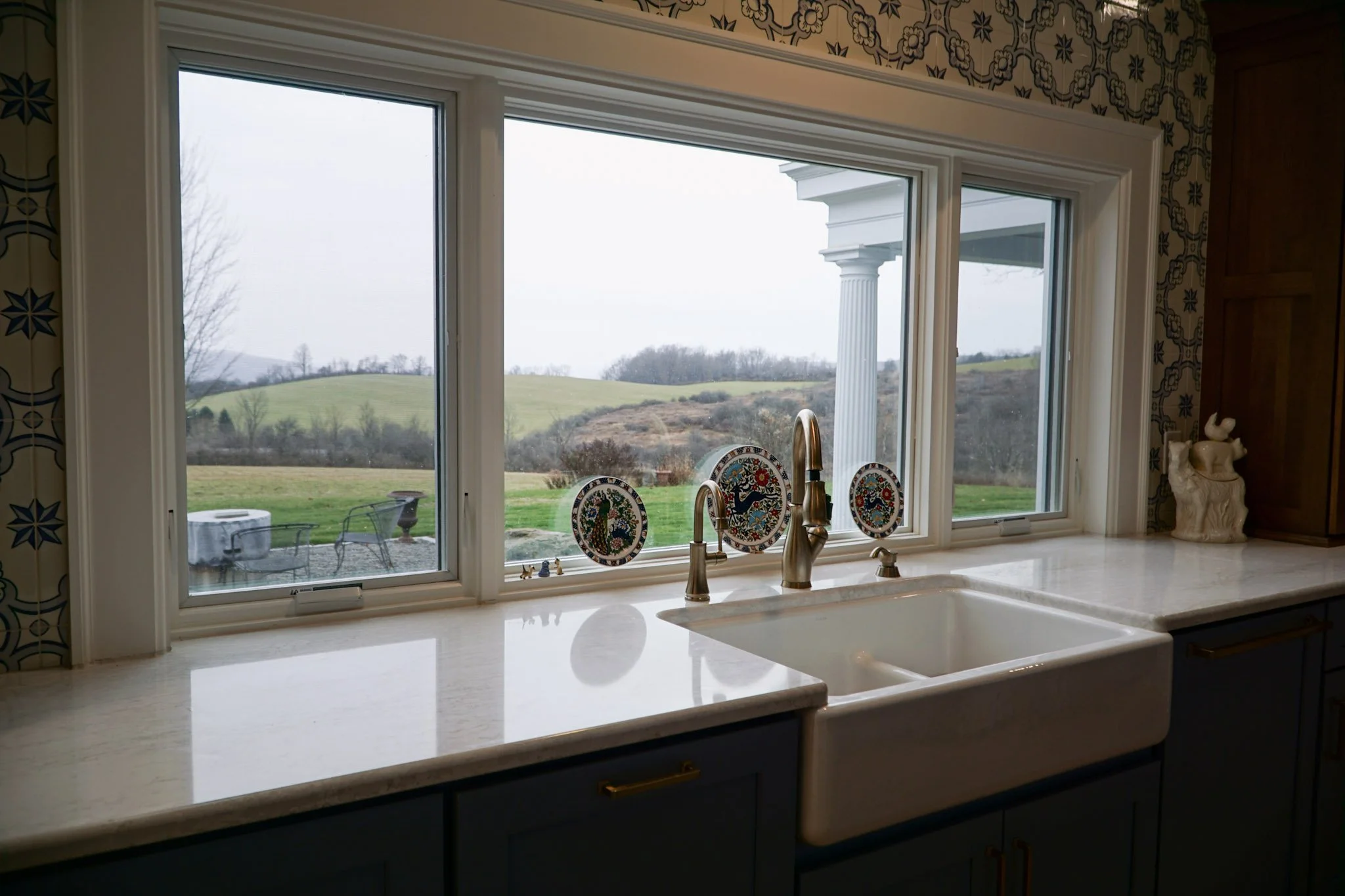 A view out a big window sectioned into three parts behind a big white ceramic sink. Three decorative plates sit along the window and blue and white tile surrounds them. A small white ceramic figure sits to the right of the sink. 