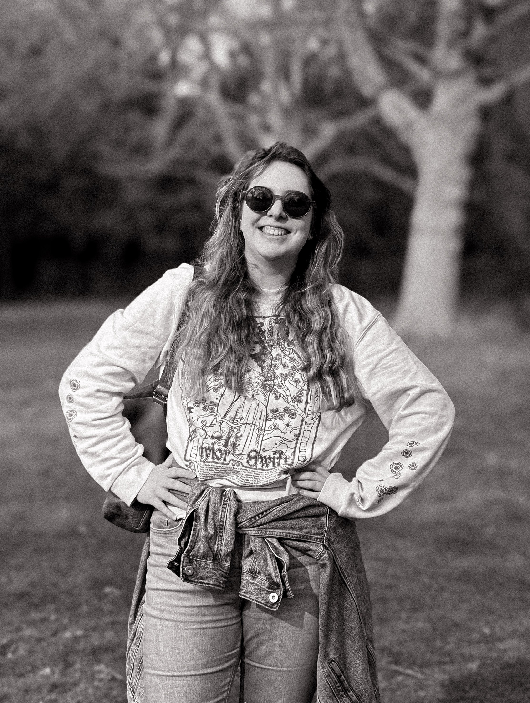 Camilla, office manager, is smiling outside in front of a few large trees with long wavy hair, sunglasses, wearing a Taylor Swift sweatshirt, with hands on hips.