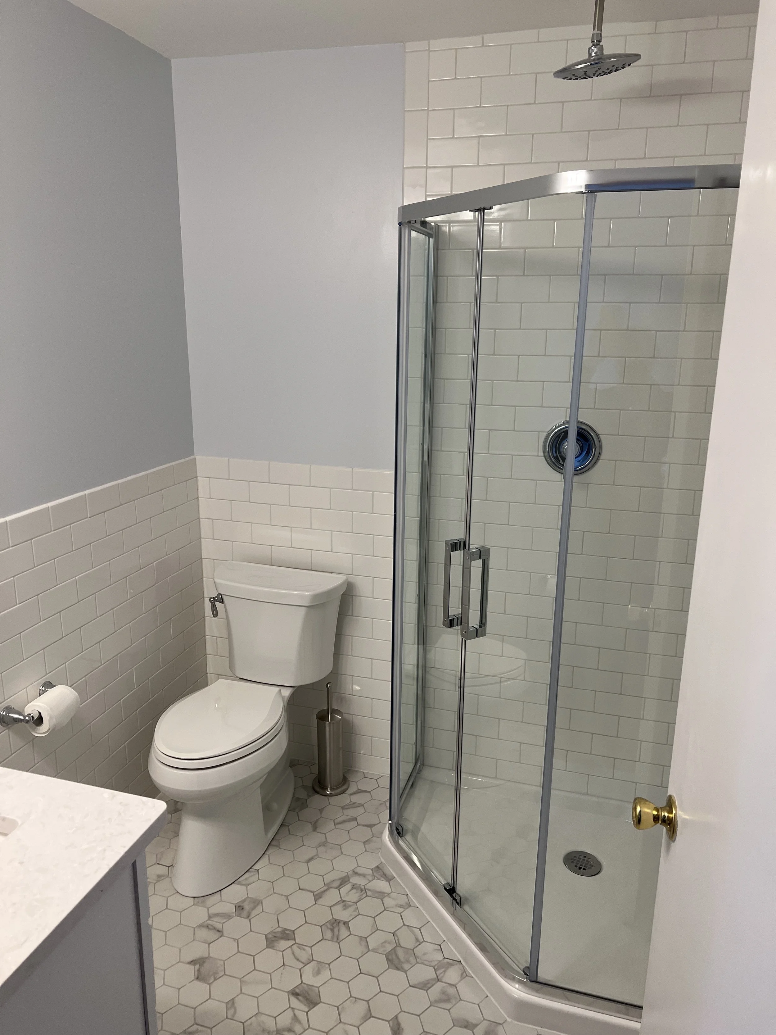 Bathroom with a toilet, a glass shower enclosure, white hexagon floor tiles, tiled walls, a ceiling shower head, and a door with a brass knob.