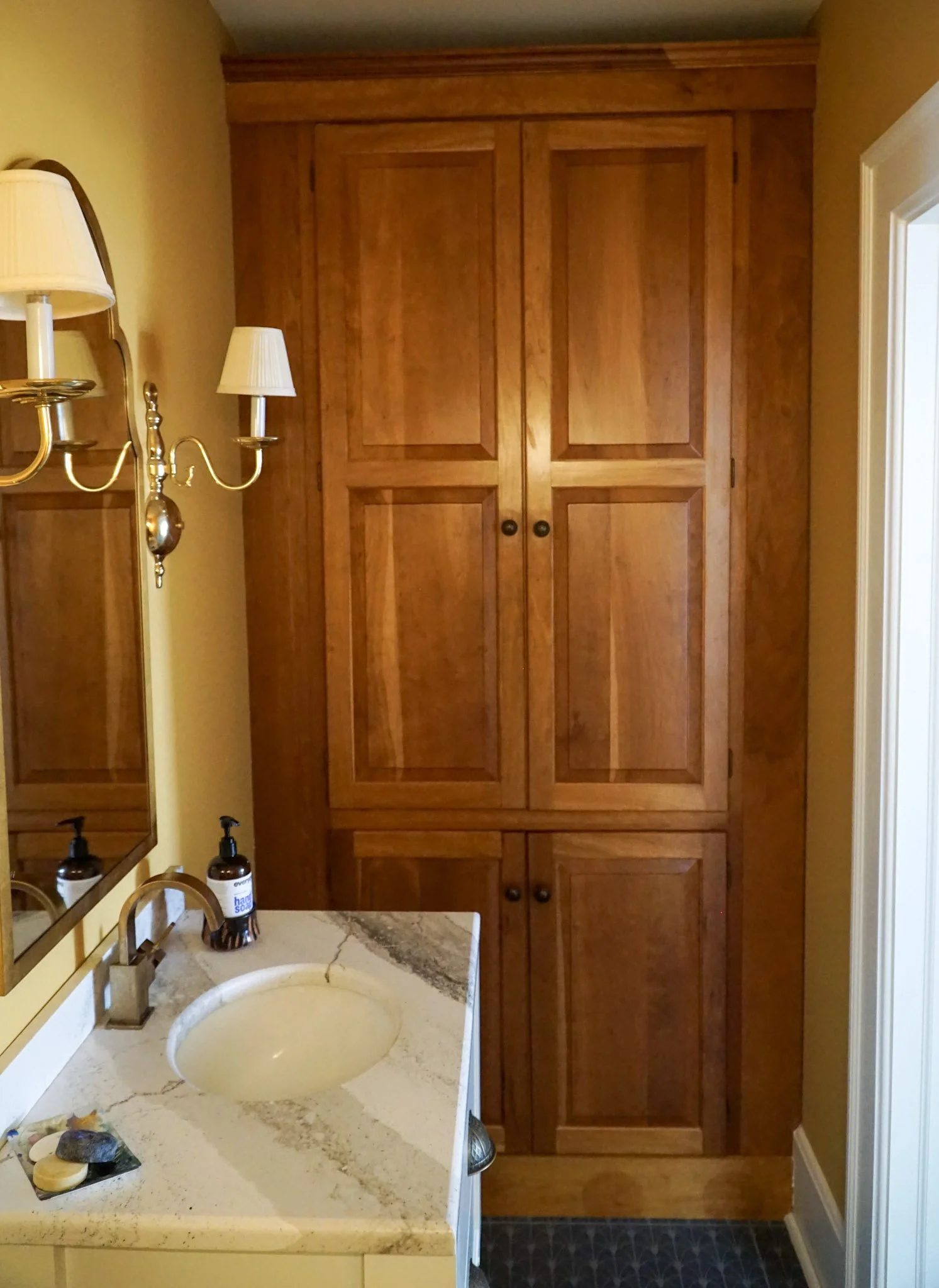 A repurposed old kitchen pantry sits at the end of a small bathroom. A marble sink with soap sits before it with a mirror and two light fixtures framing it. The wood of the old pantry is a warm oak and there are blue tiles for the floor. 