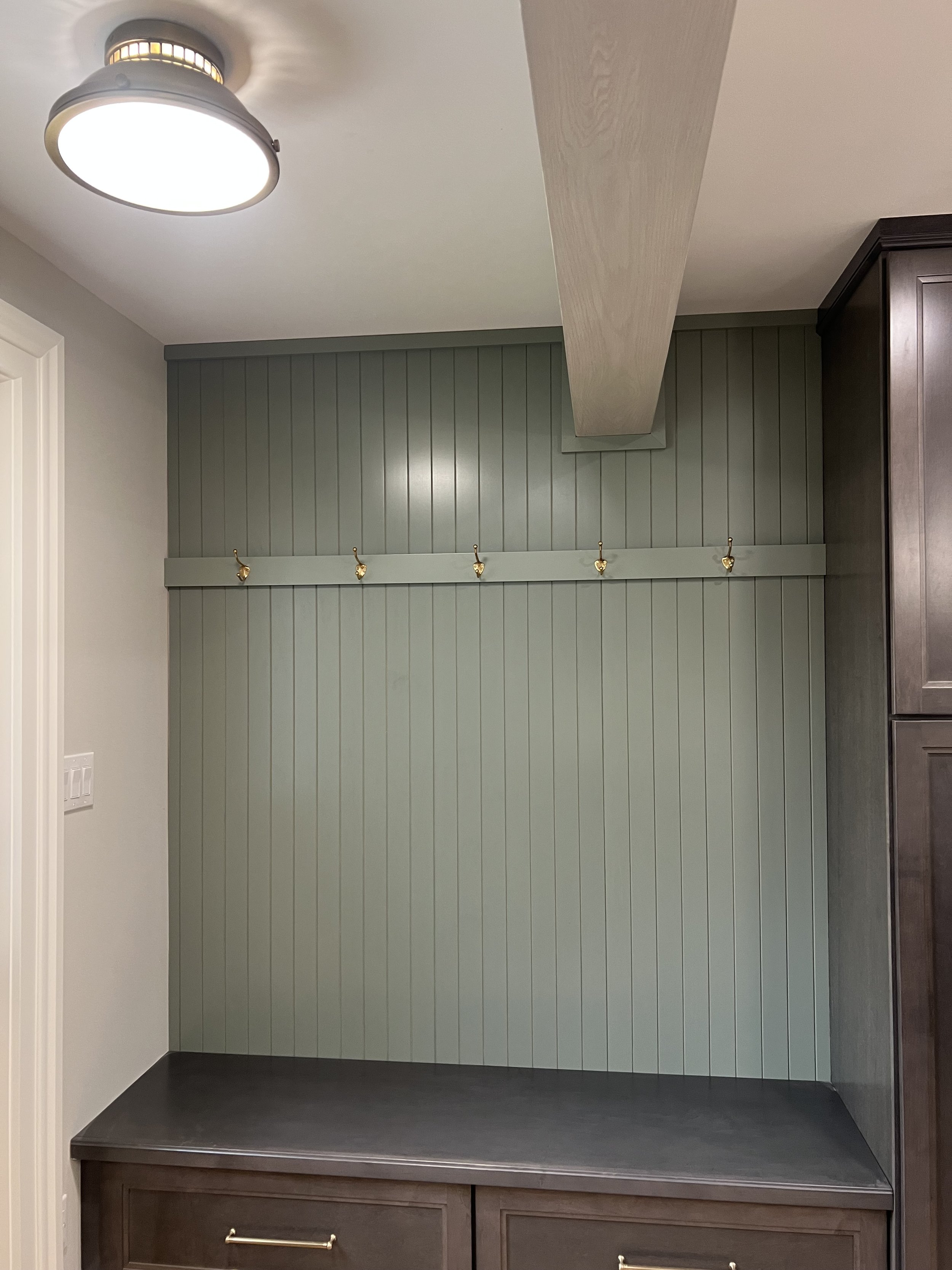 This mudroom drop zone with sage green bead board, brass hooks and maple cabinetry provide storage near the entrance which is located in a newly renovated kitchen in Central New York.