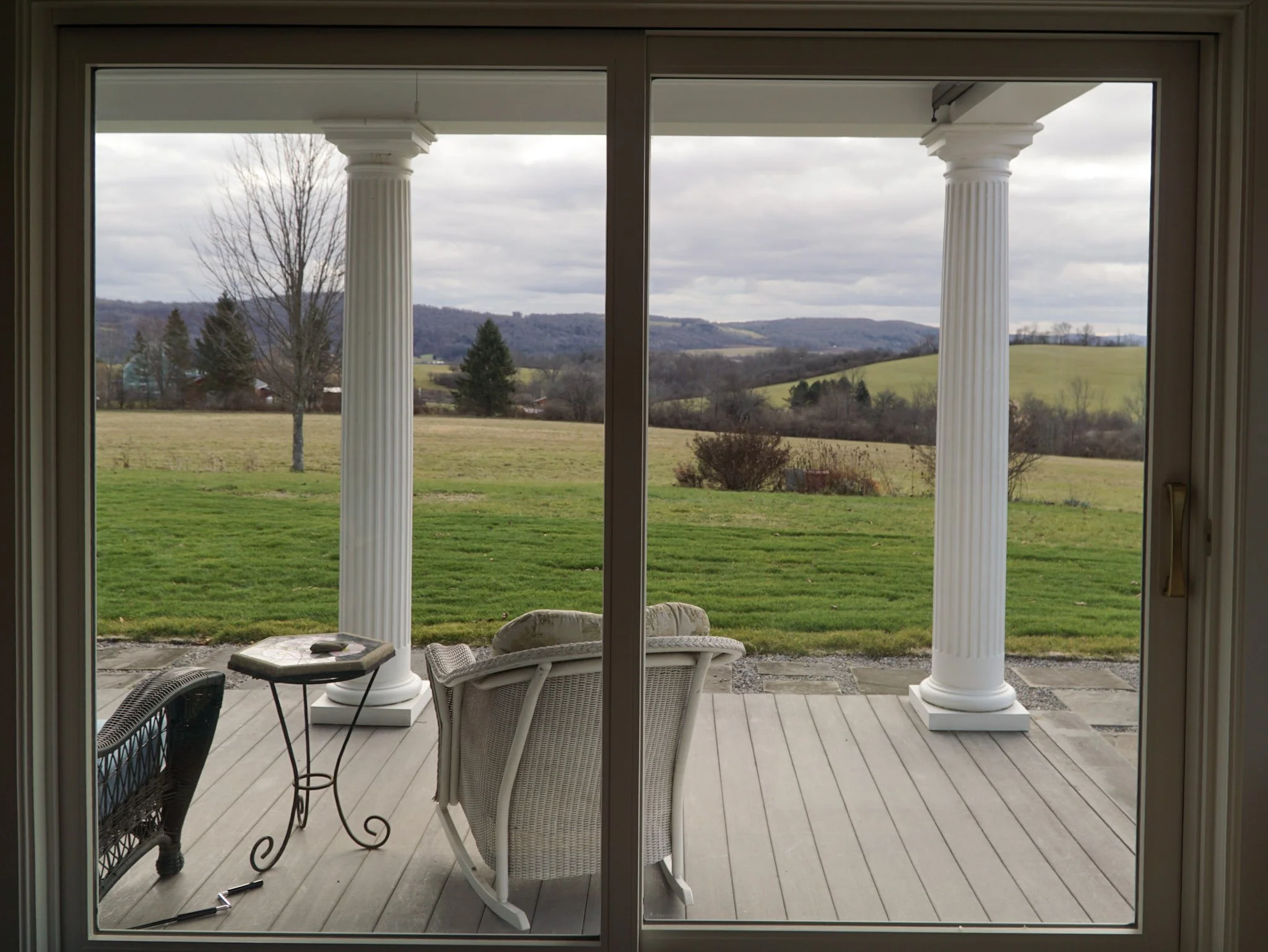 A big sliding glass door looks out onto the view of a lawn that leads into a pasture and rolling hills. Two pillars indicate a porch area with a rocking chair and table visible. 