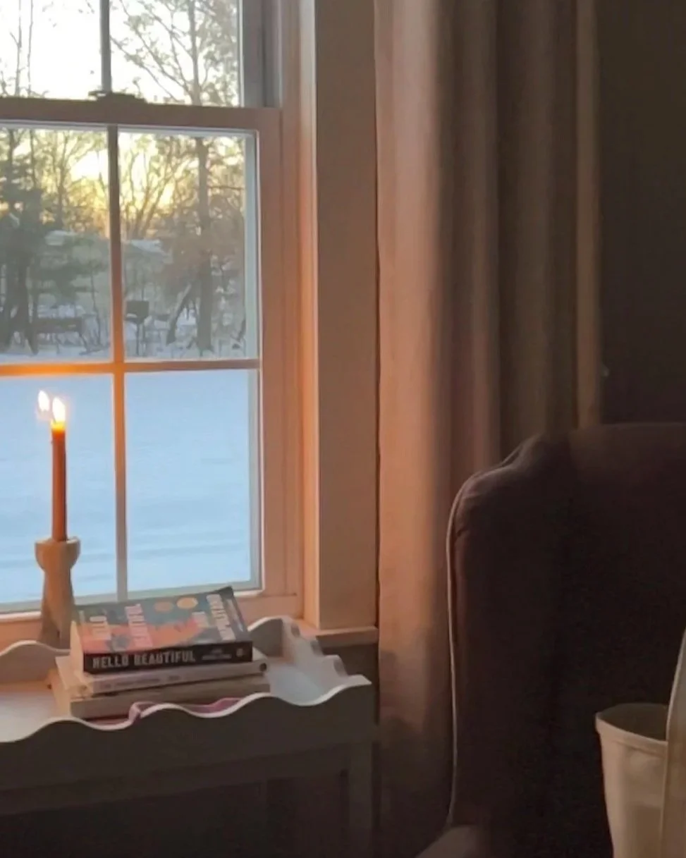 A candle sits lit in a window next to a blue end table with a stack of books upon it. A soft mauve colored chair sits next to the table in this central new york home.