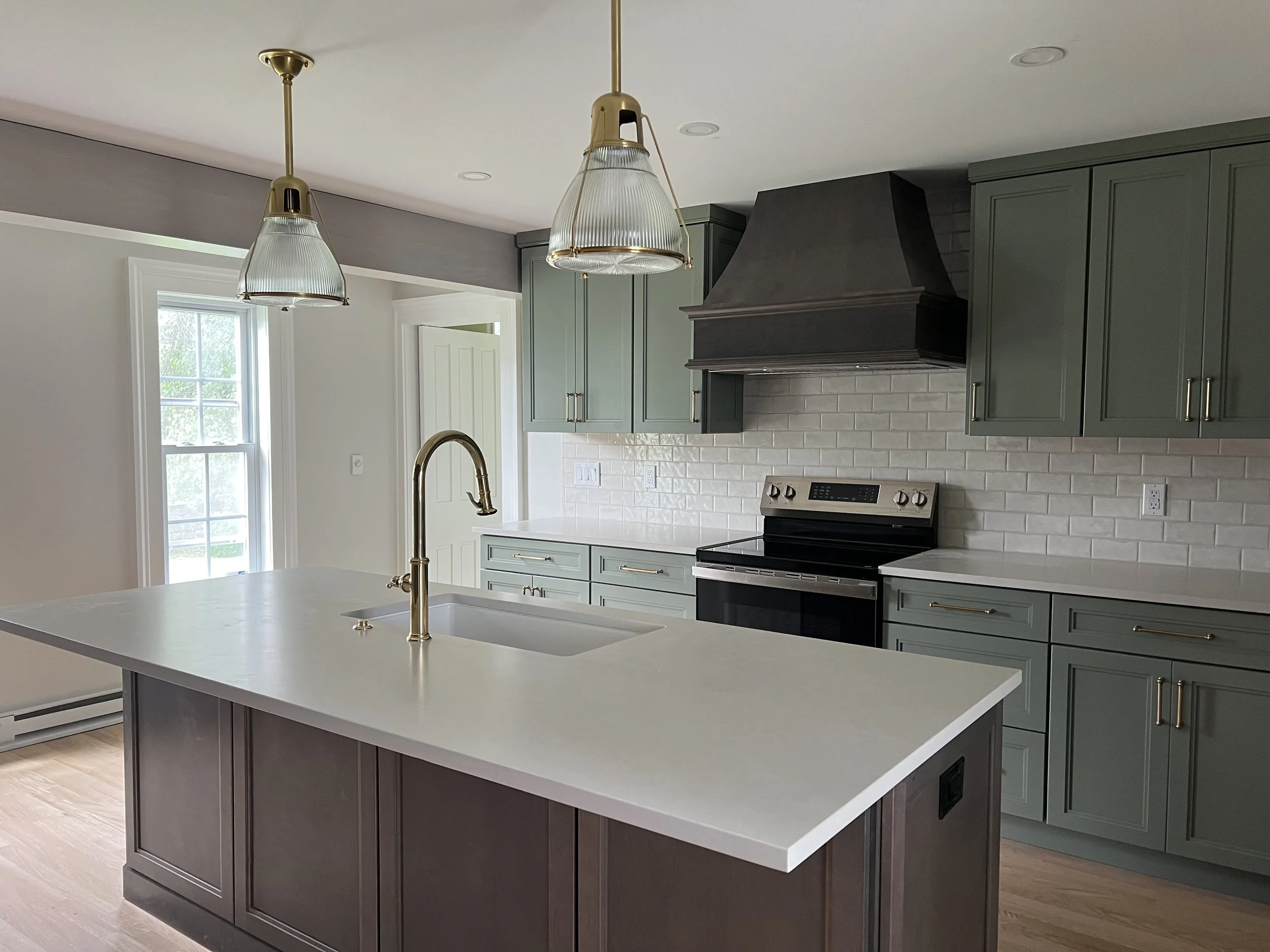 A new kitchen renovation completed by a custom home builder in Hamilton, NY. Light sage green cabinets with a stained maple island and stained maple range hood, brass fixtures, an unlaquered brass faucet. 