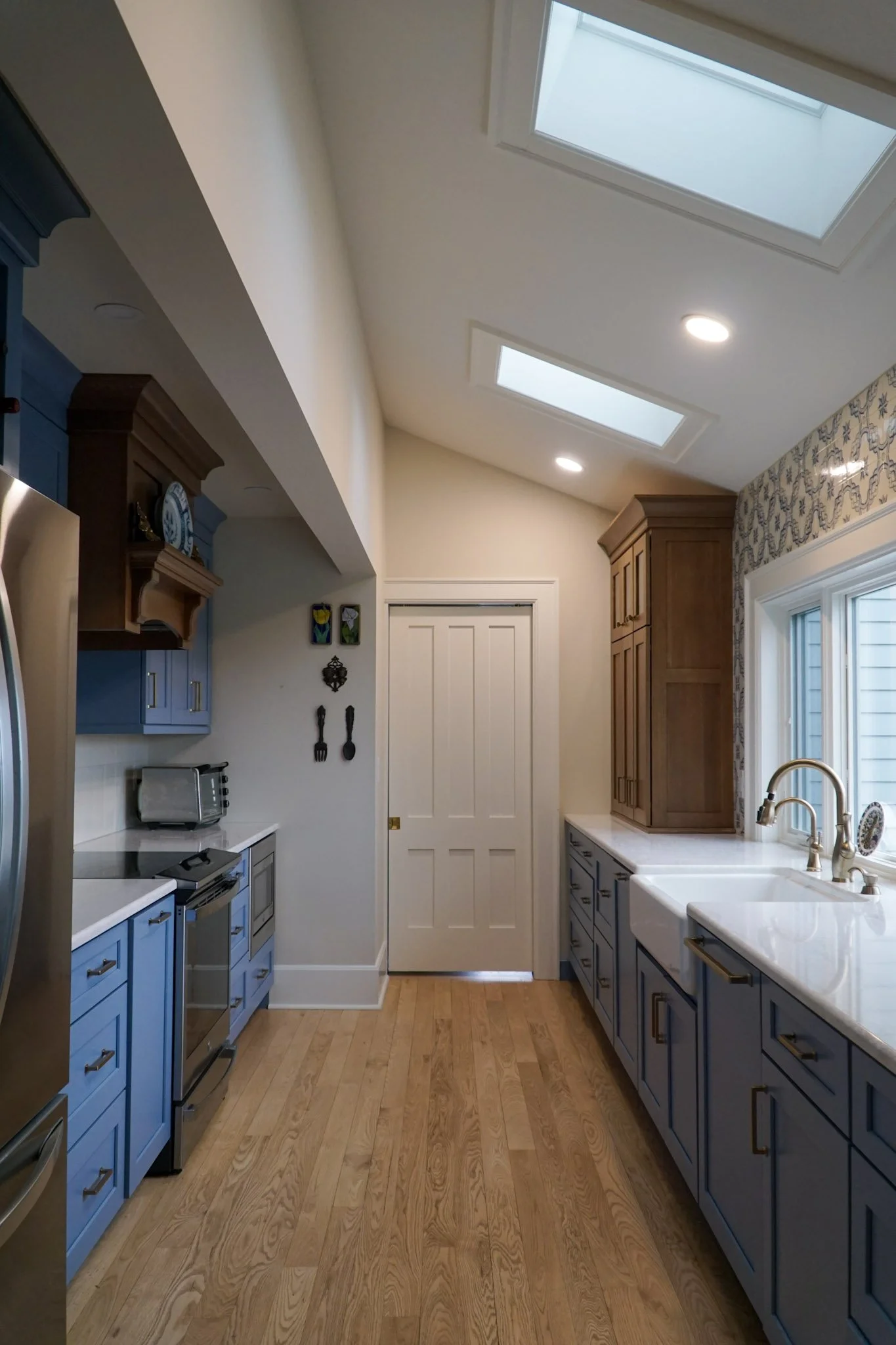 A galley kitchen with semi-custom blue and wooden cabinets. Hardwood floor with a white modern sink, two skylights and a door at the end of the kitchen. 