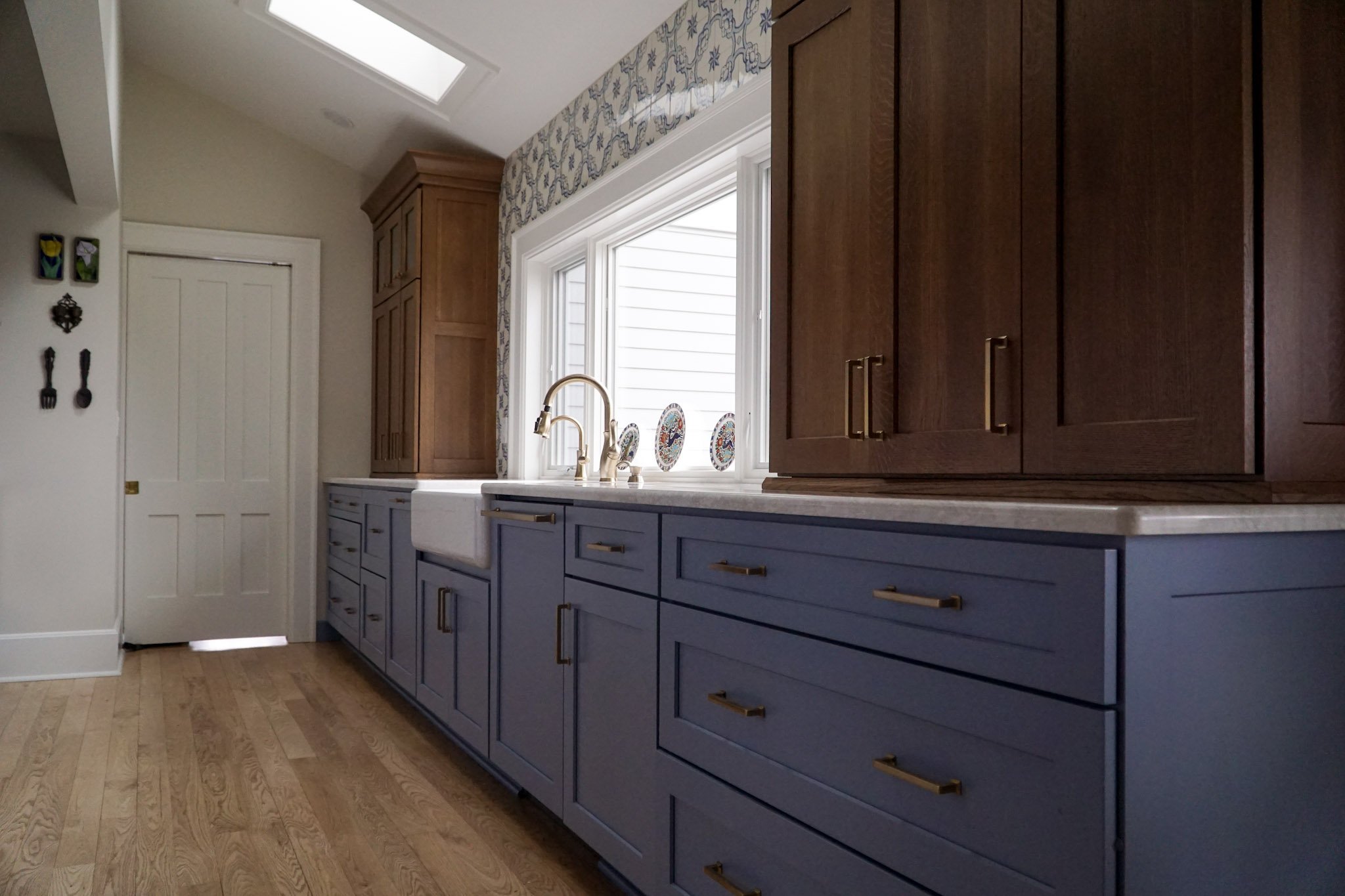 Blue semi-custom cabinets line the bottom with white marble counter tops. Resting on these countertops are some wooden cabinets for extra storage. A big window sits behind the sink and hardwood flooring is present. 