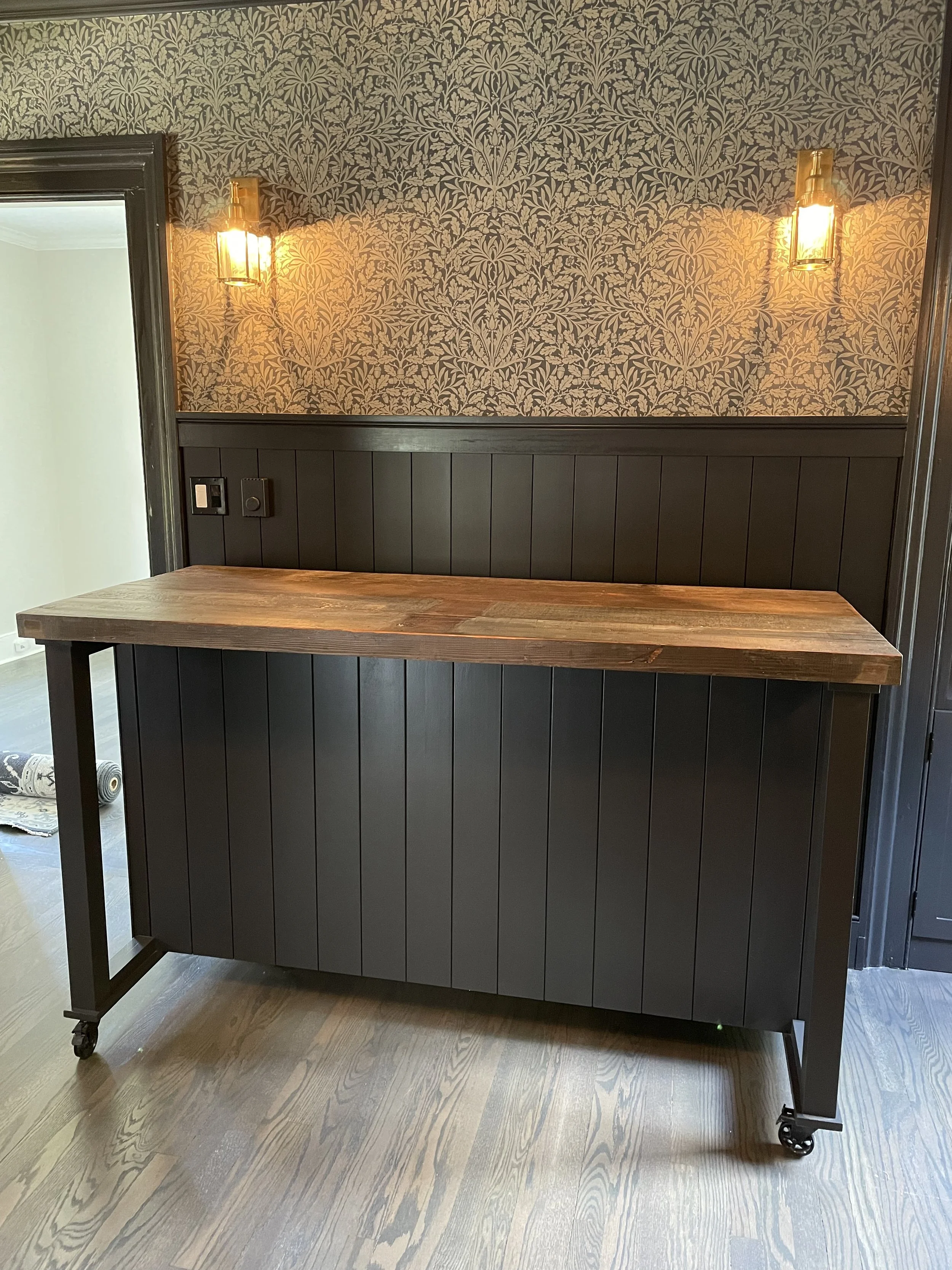 A black kitchen island with a wooden top against a wallpapered wall with vanity lights.