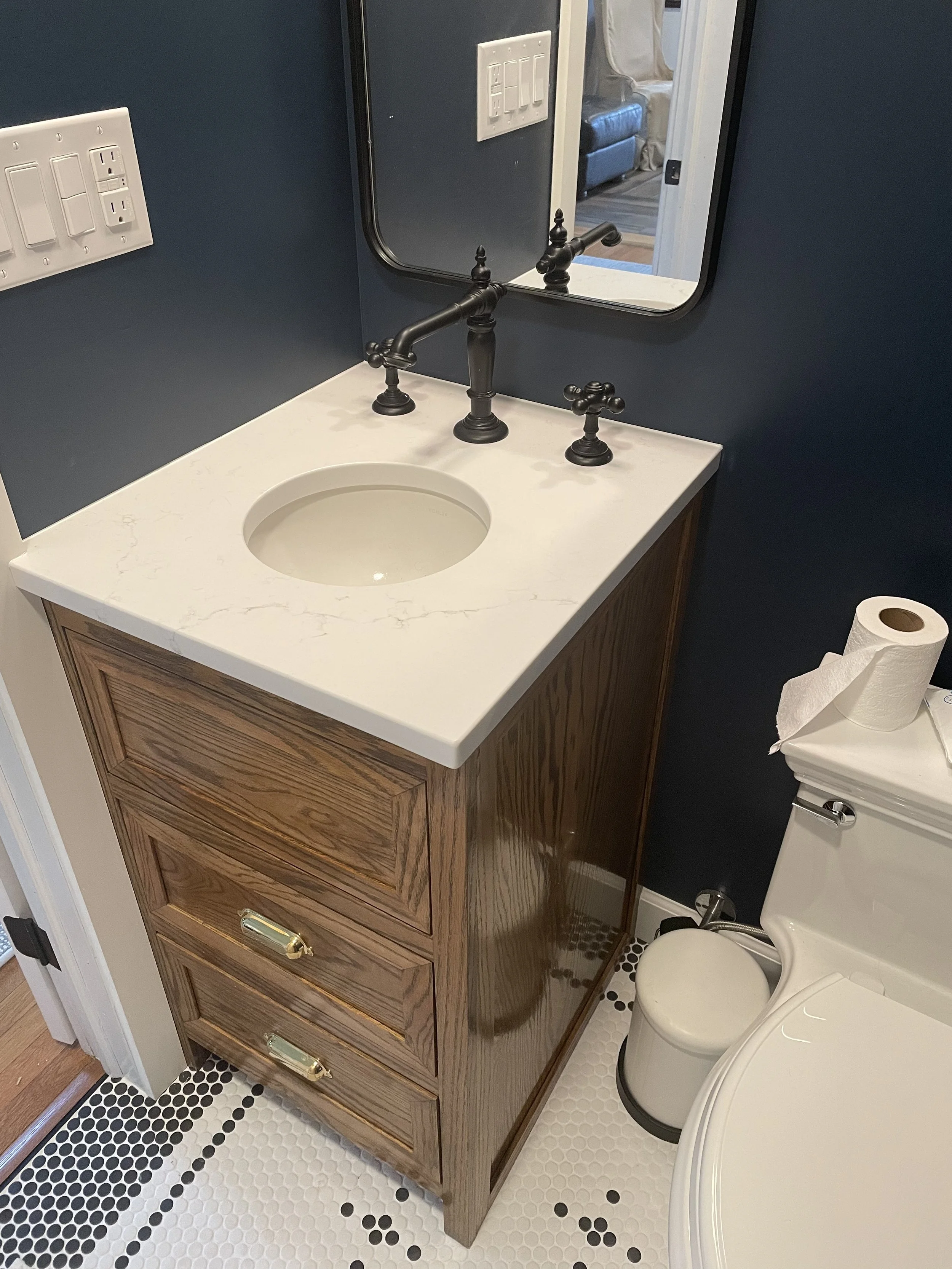Bathroom vanity with a white marble countertop, a circular sink, and black faucet fixtures. A mirror above the sink, with a dark blue wall behind. To the right, a toilet with a roll of toilet paper on top.