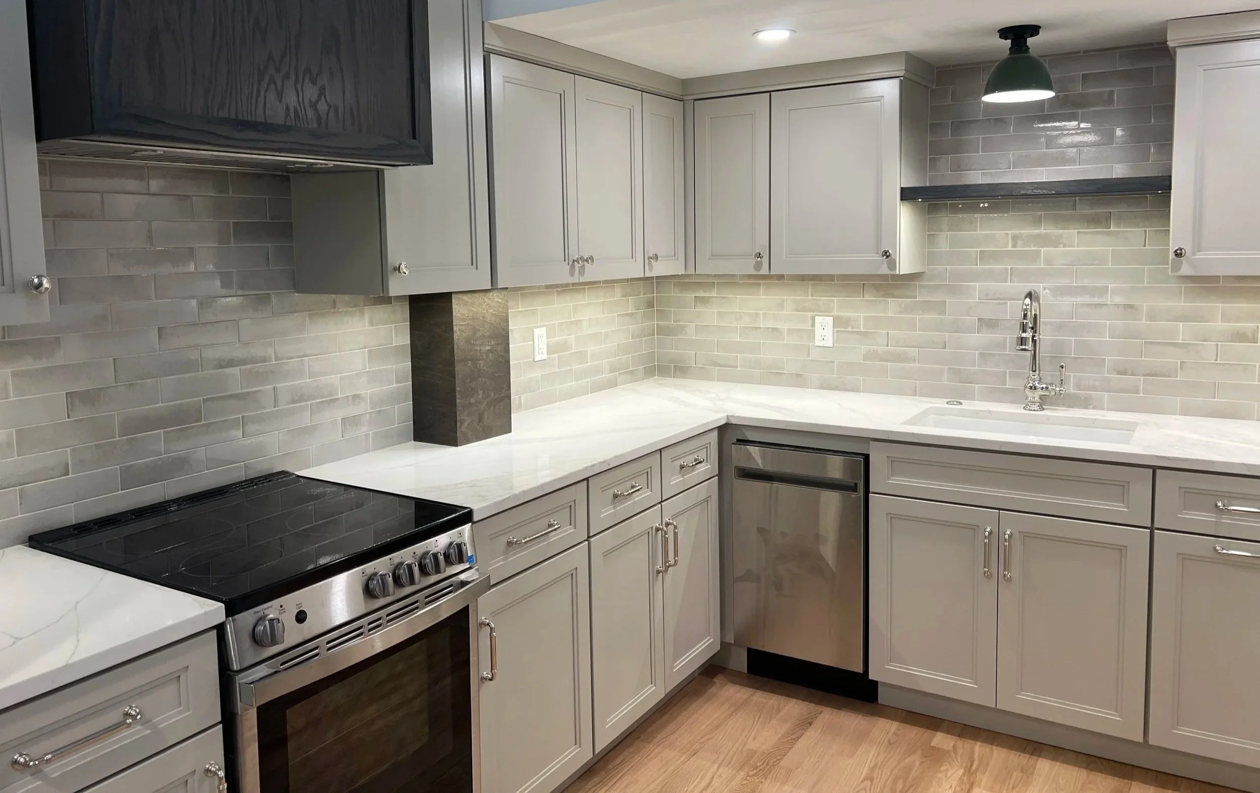 A small kitchen with soft gray cabinets, white quartz countertops, a stained maple range hood, gray subway tile backsplash, stainless steel appliances and nickel sink faucet. Part of a kitchen renovation in Hamilton, NY.