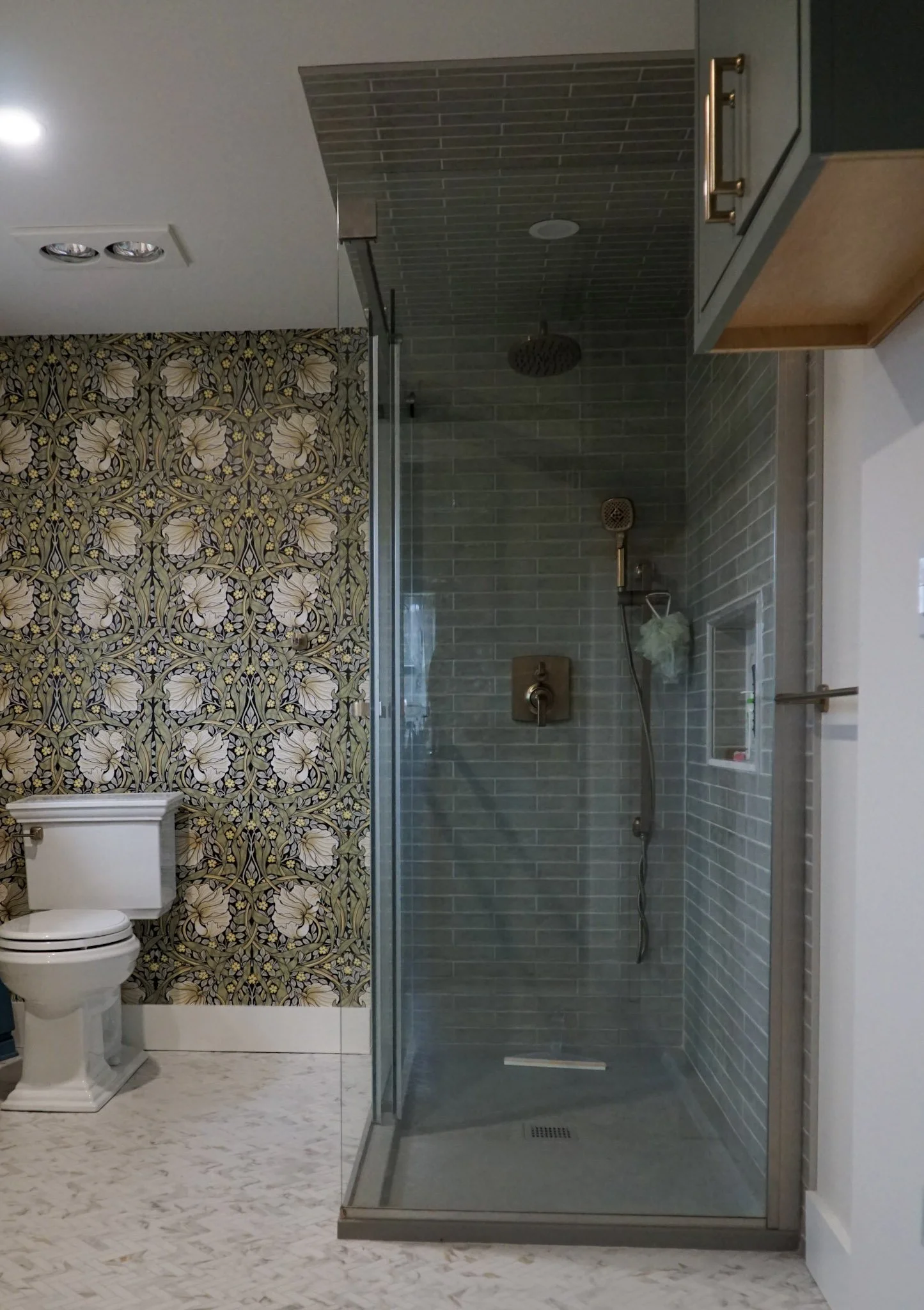 A modern step-in shower with gray subway tiling sits on the right side of the photo with a toilet on the left. Dramatic wallpaper and a green floating cabinet can be seen. White tile flooring