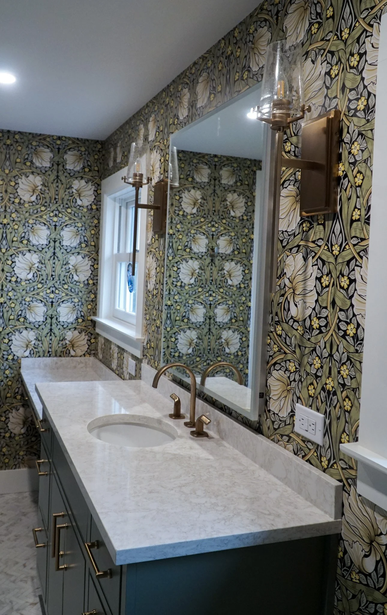 A marble sink top in front of a mirror with two light fixtures on either side. Green cabinet beneath the sink and a dramatic green wallpaper with white flowers and black accents. A window at the end.