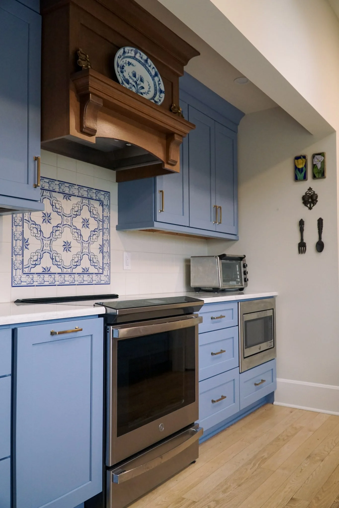 Semi-custom blue cabinets on display with a wooden stove hood and modern oven. intricate blue and white tile backdrop behind the stove top. A microwave and toaster are in the back as well as a few things hanging on the wall. 