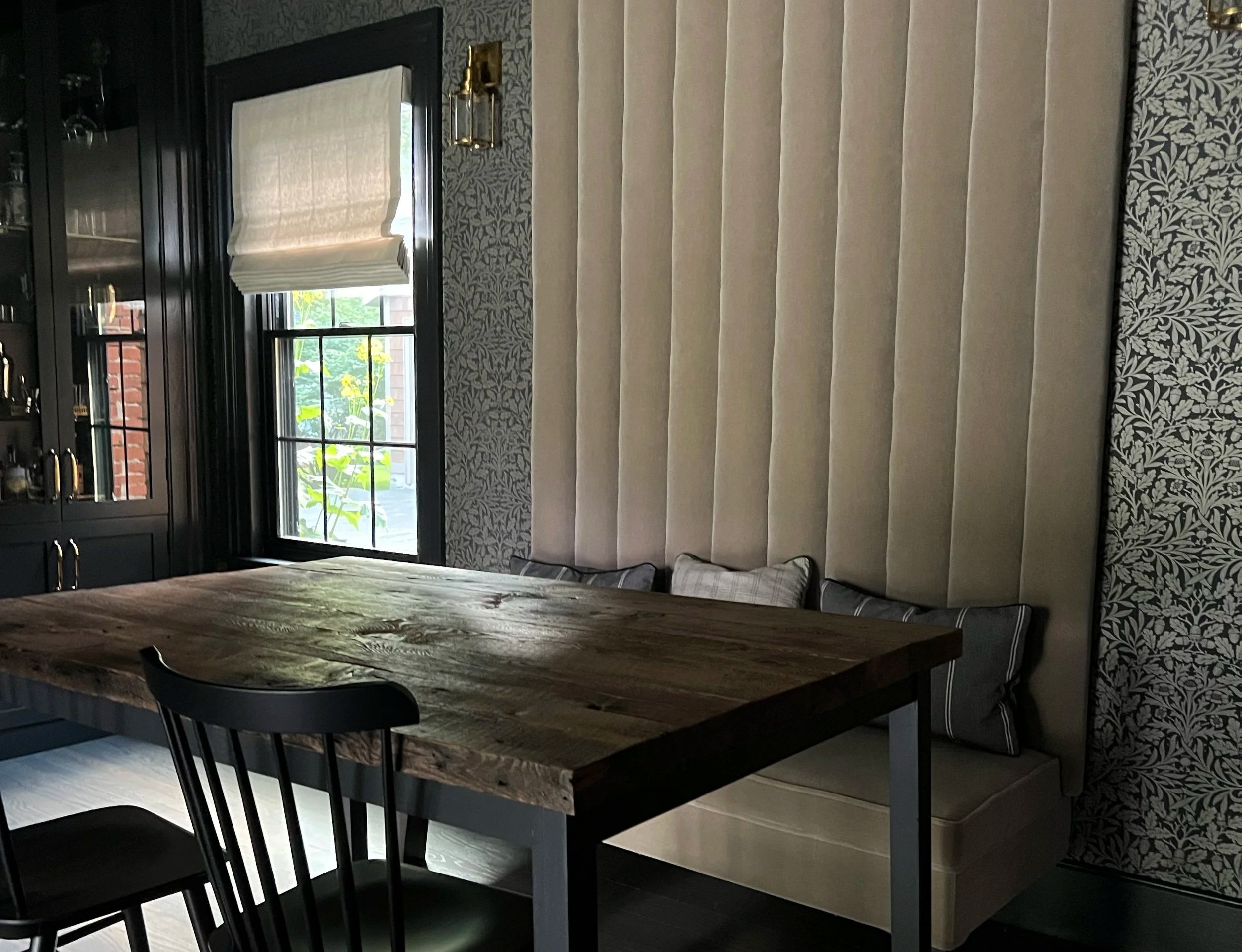 A dining room table sits in front of an extensive seating area. Wallpapered walls and a cabinet sits at the left edge of the frame.