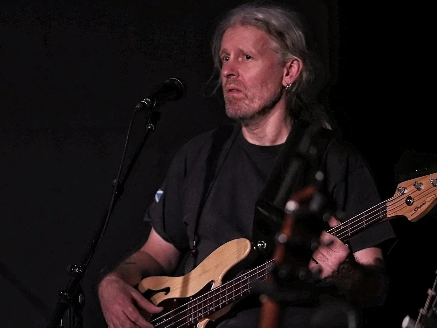 Man houdt een akoestische gitaar voor zijn gezicht terwijl hij direct naar de camera kijkt, met een bakstenen muur op de achtergrond.