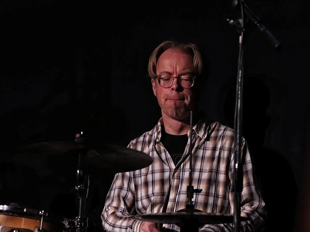Man met lang haar, bril en een zwart shirt, met twee drumstokjes voor zijn gezicht, tegen een bakstenen muur.