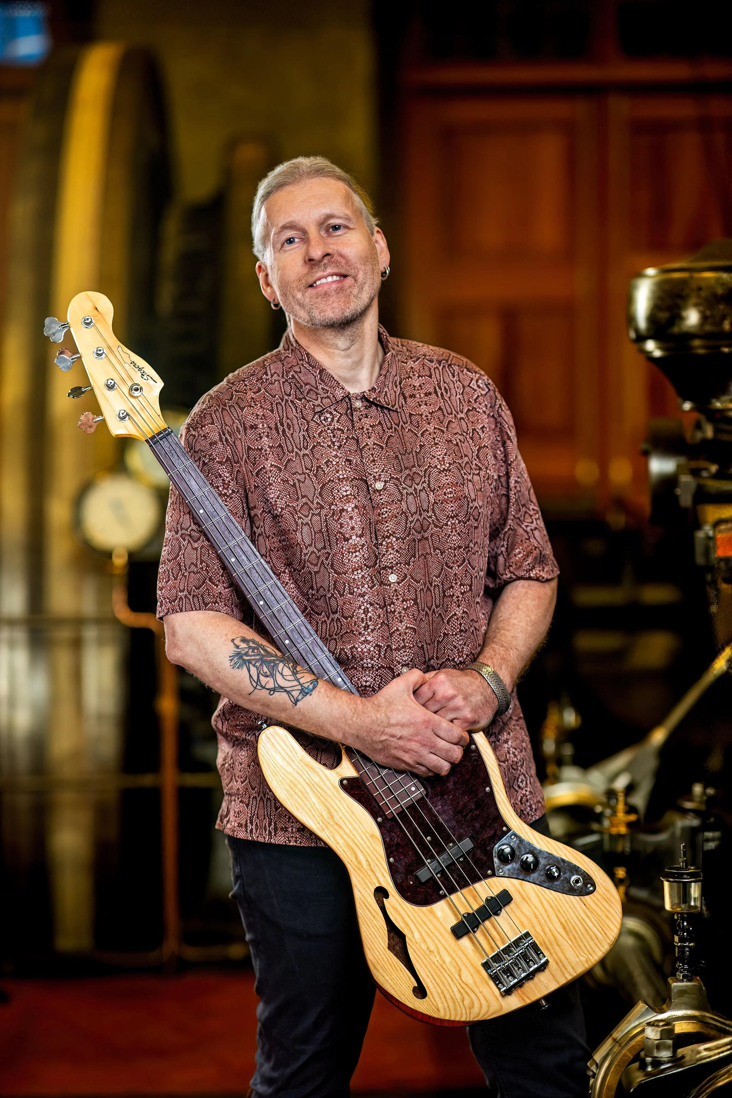 Man houdt een akoestische gitaar voor zijn gezicht terwijl hij direct naar de camera kijkt, met een bakstenen muur op de achtergrond.
