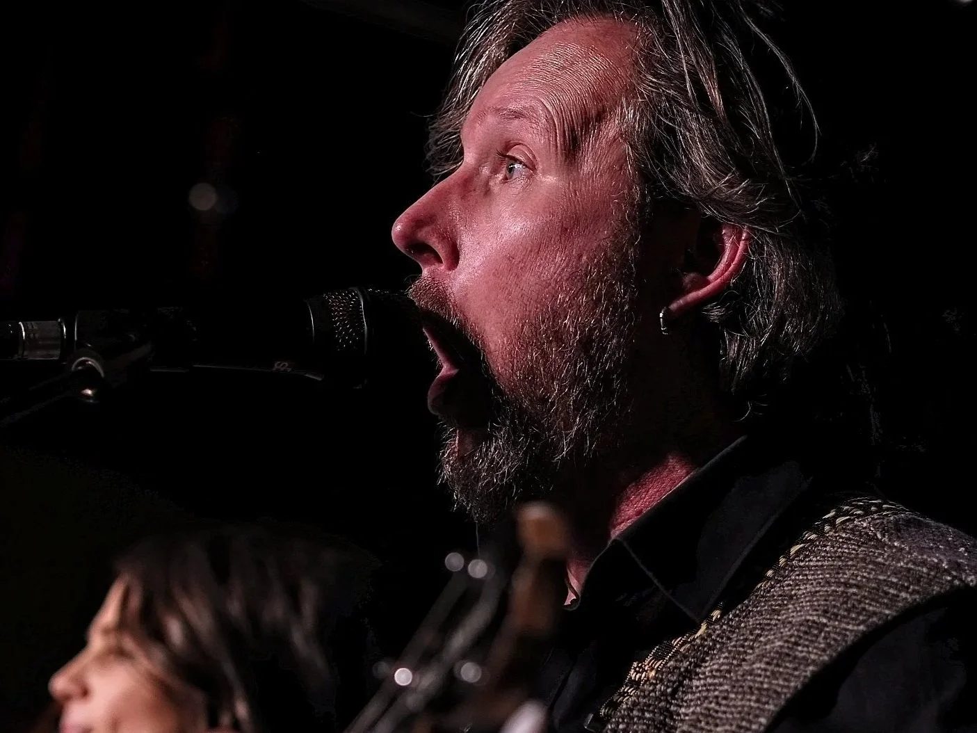 Man met een baard en lang haar houdt een gitaar vast dicht bij zijn gezicht, met een bakstenen muur op de achtergrond.