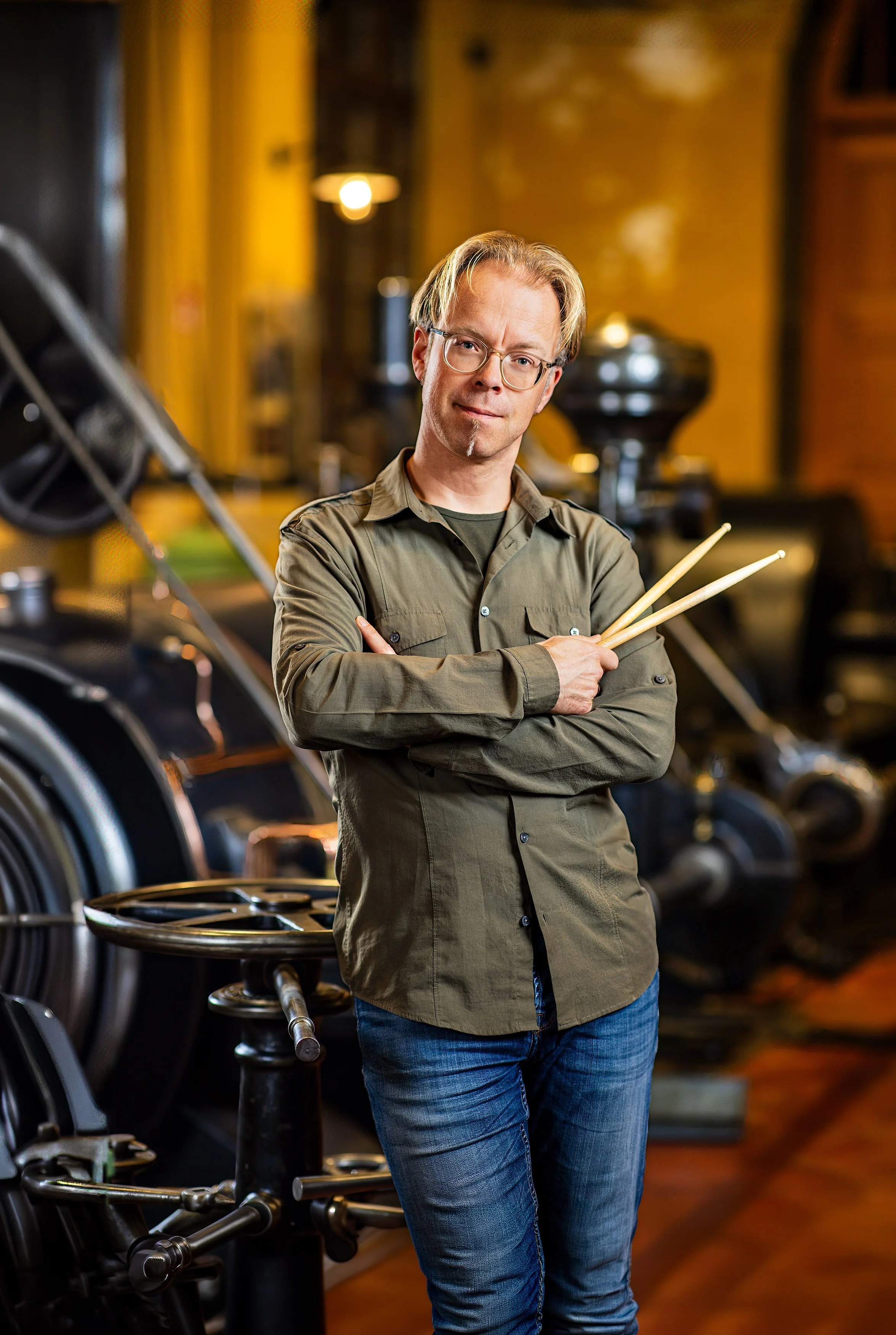 Man met lang haar, bril en een zwart shirt, met twee drumstokjes voor zijn gezicht, tegen een bakstenen muur.