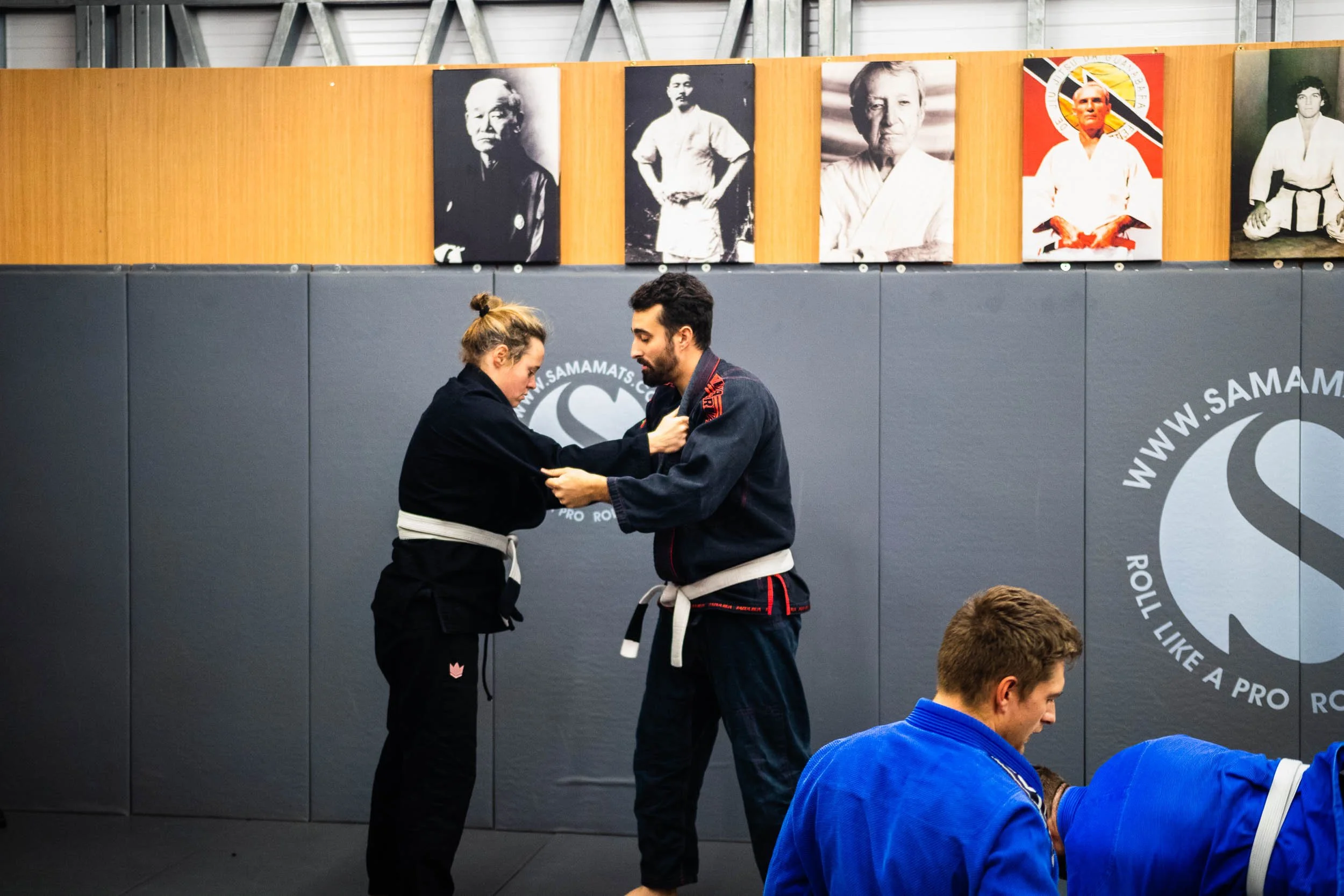 Two martial arts students practicing Brazilian Jiu-Jitsu with a woman adjusting the man's gi, in a training gym with images of martial arts legends on the wall.