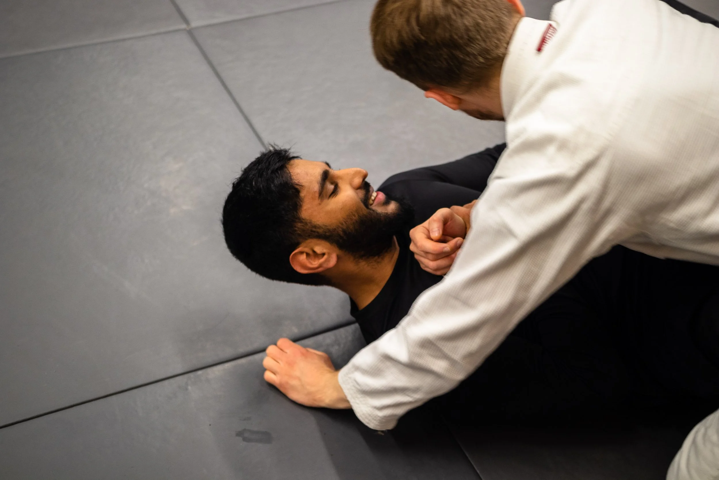 Two men practicing Brazilian jiu-jitsu, one lying on the ground with a smile and the other kneeling over him, gripping his collar.