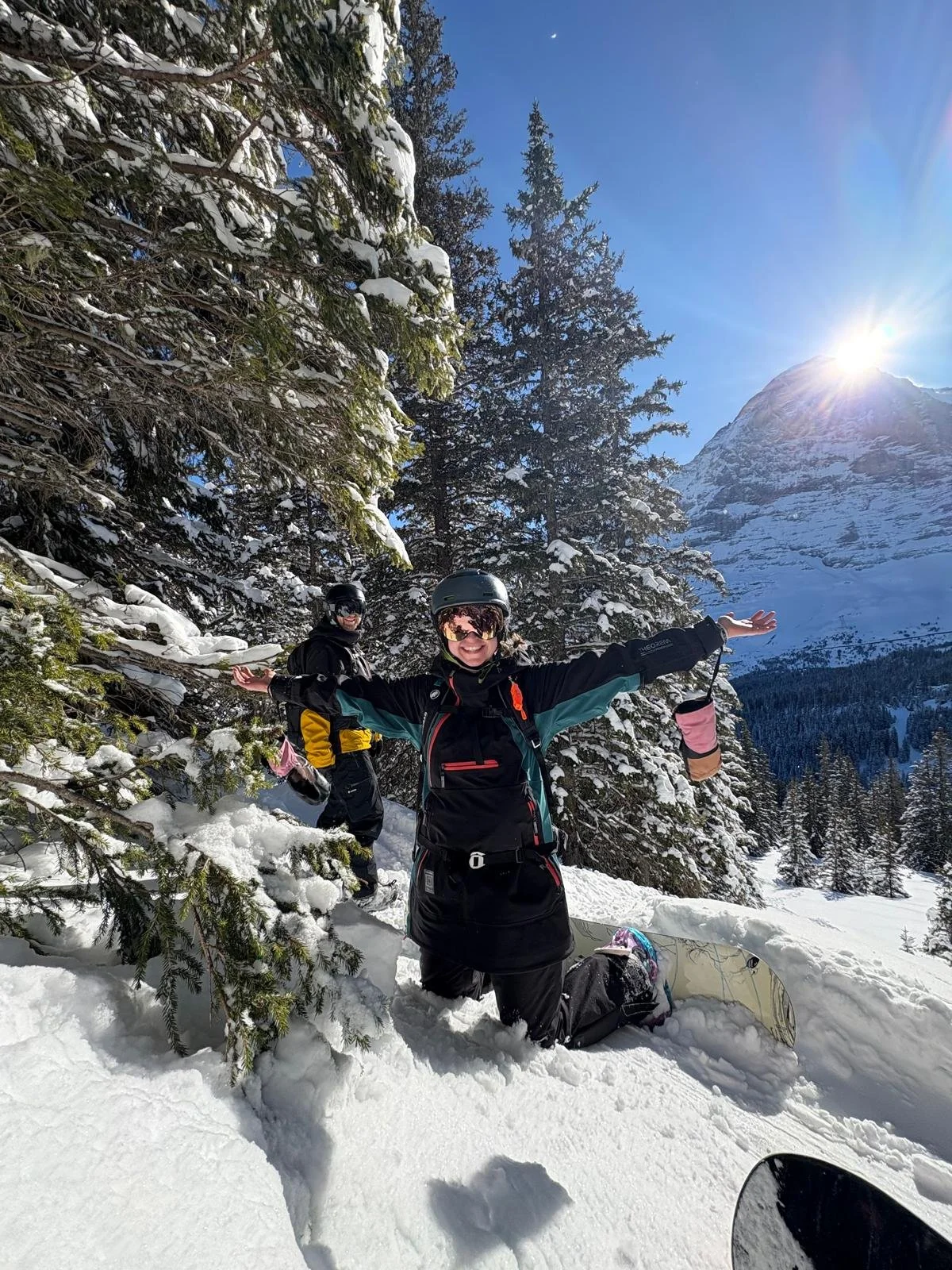 Kundin Ilse lächelt mit Snowboard im Tiefschnee, Arme ausgebreitet, im Hintergrund schneebedeckte Berggipfel im Gegenlicht