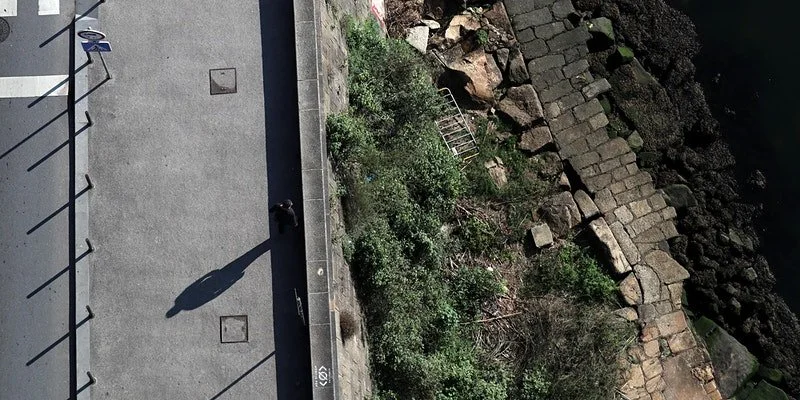 Left hand side of image is footpath with shadow of person walking. Right hand side is marginal shrubby land with rubbish and old wall.