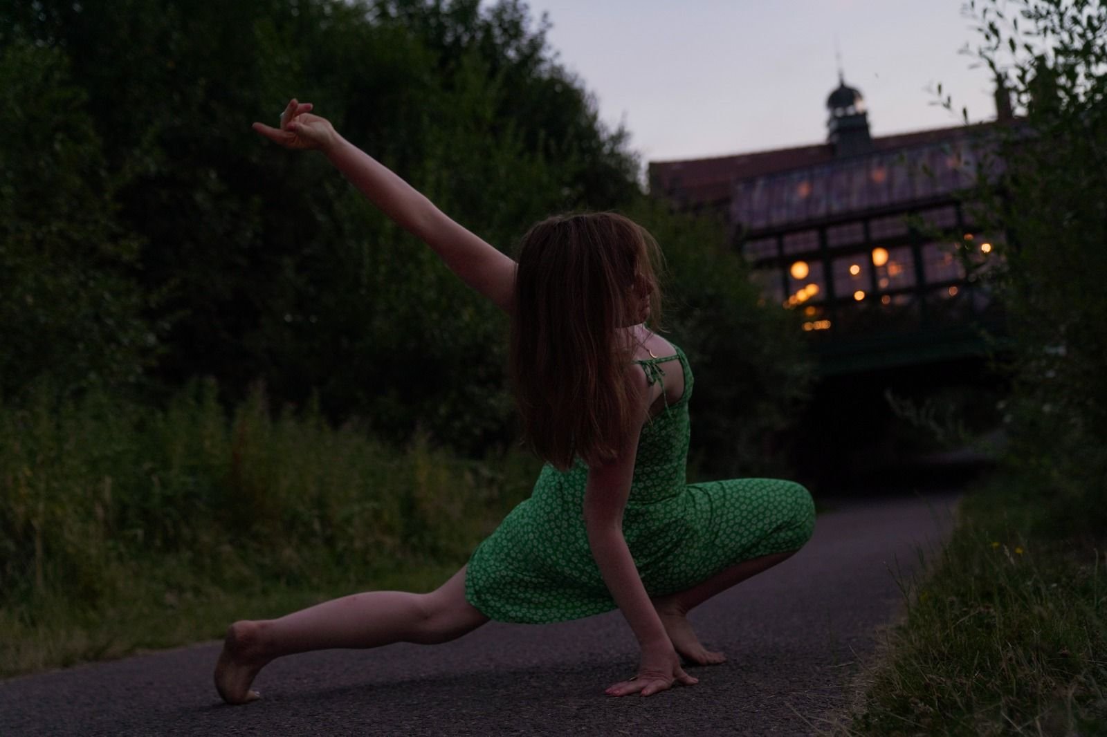 A woman practicing yoga on a paved path during dusk, wearing a green dress, balancing with her left hand and foot on the ground, her right arm extended upward, with trees and a building with lit windows in the background.
