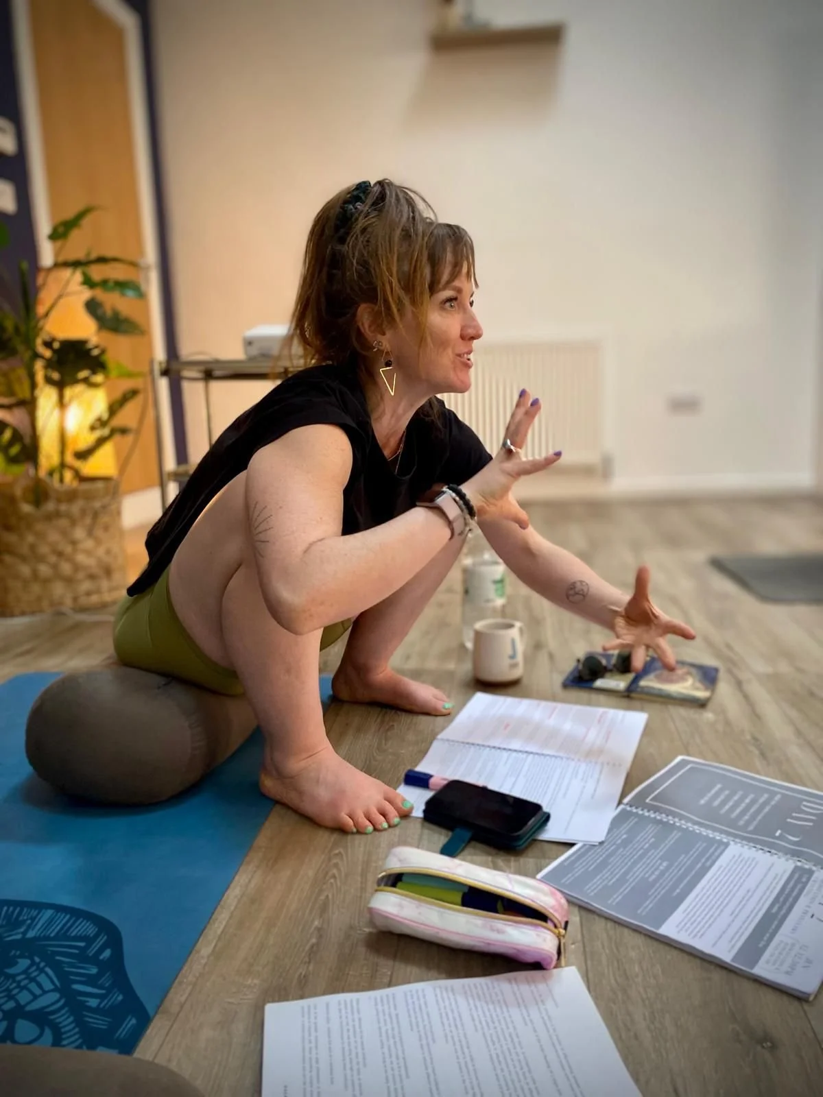 A woman practicing yoga indoors on a mat, balancing on her hands and feet with her knees bent, surrounded by papers, books, and personal items, with a plant and a radiator in the background.