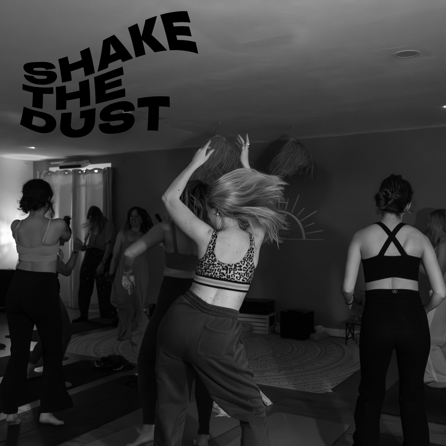Group of women dancing in a room with a wall decoration and a sunset graphic, with the text 'Shake the Dust' in bold 3D letters in the upper left corner.