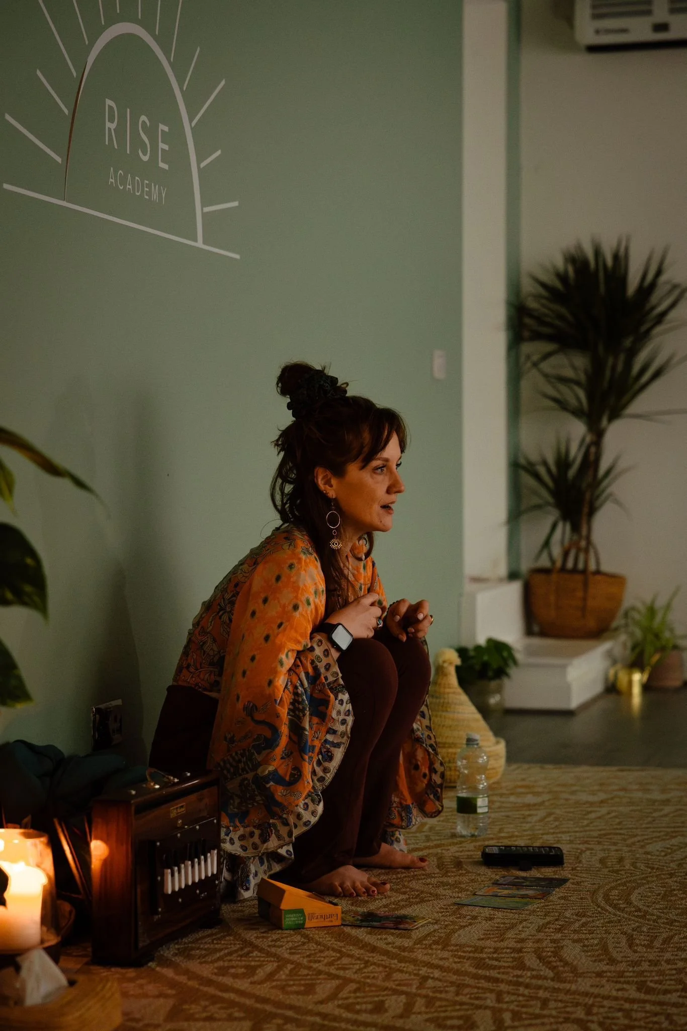 A woman with dark hair tied up in a messy bun, sitting cross-legged on a rug in a room with green walls. She is wearing a colorful, patterned top and brown pants, and appears to be speaking or giving a class. There is a sign on the wall that reads 'RISE ACADEMY' and some plants and objects around her, including a water bottle, a smartphone, and tarot cards.
