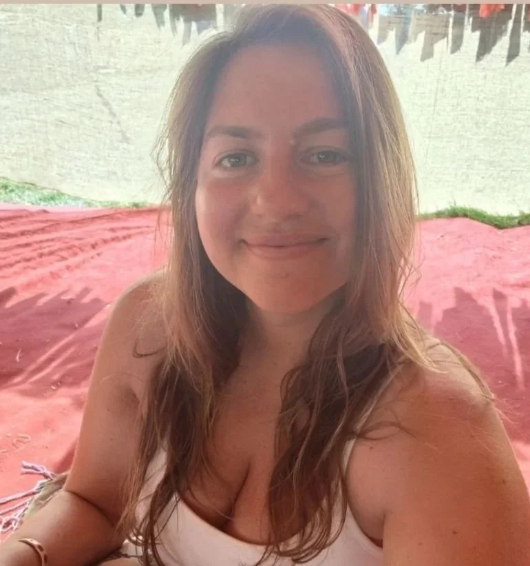 A young woman with long, wavy brown hair smiling, sitting on a pink blanket outdoors with grass and a white wall in the background.