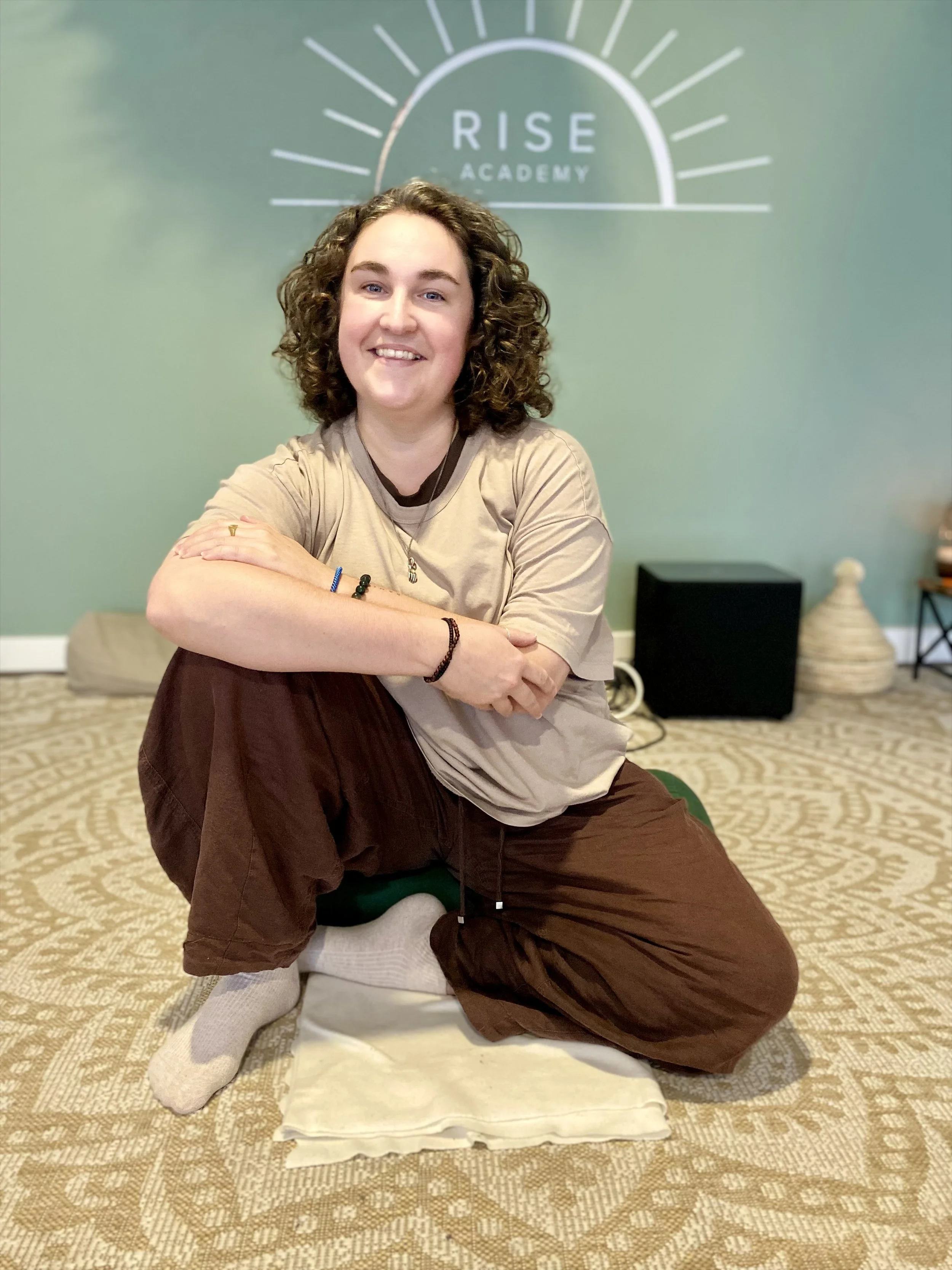 Smiling woman sitting cross-legged on a cushion in front of a wall displaying the “RISE ACADEMY” logo.