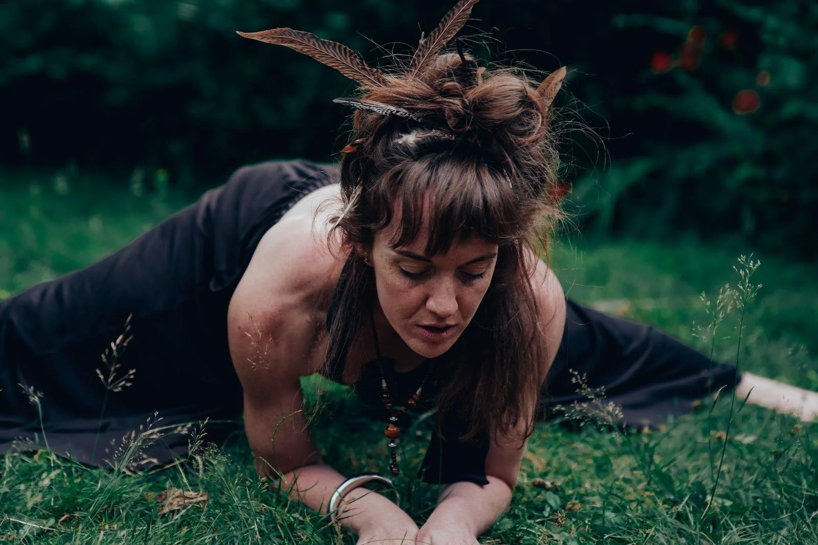 A woman with long brown hair, adorned with feathers, is lying on the grass outdoors with her eyes closed, appearing to be in a contemplative or meditative state.