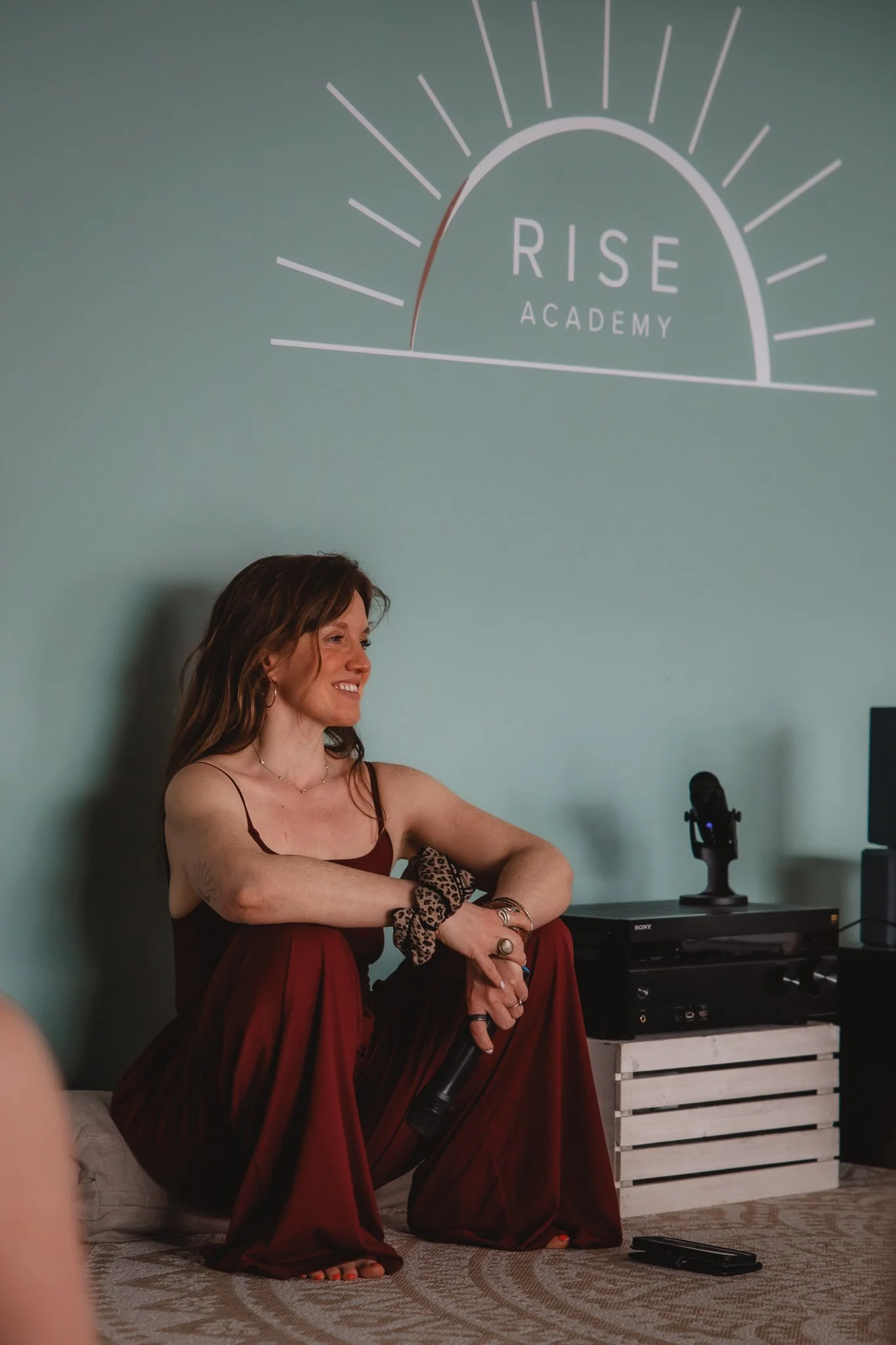 A woman with red hair holds a microphone close to her mouth with one hand and raises her arm in front of a teal wall. She wears a sleeveless dress with a leopard print patch near her waist and multiple bracelets on her wrist.