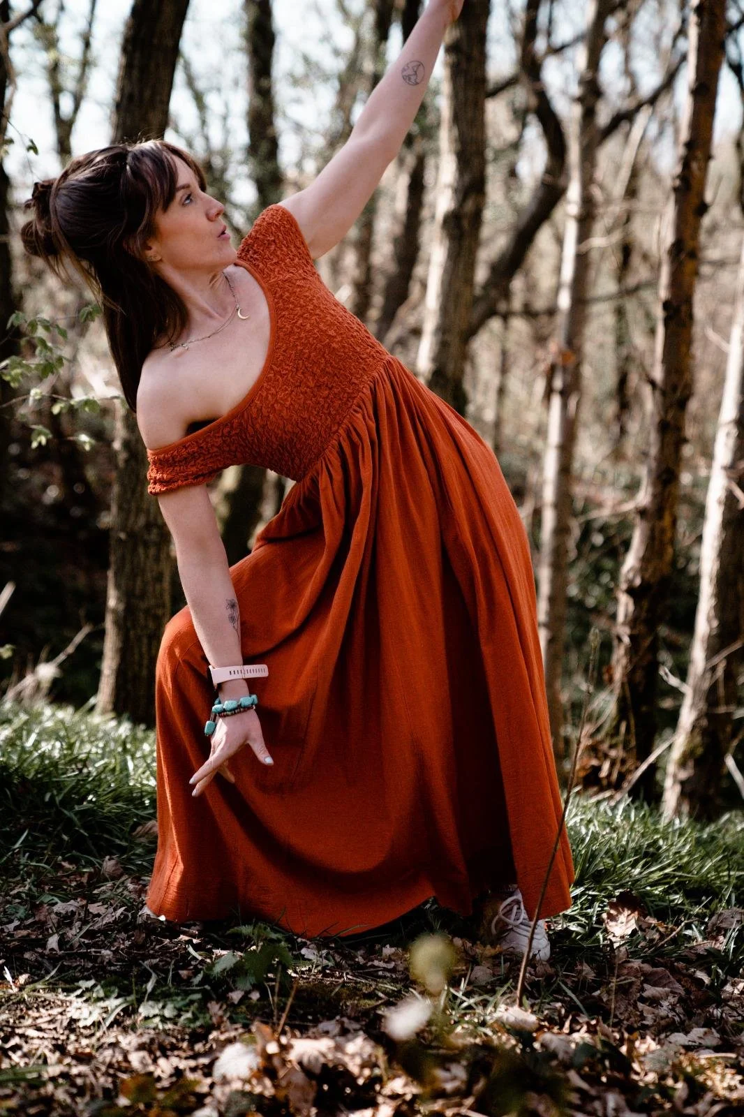 A woman in a burnt orange off-shoulder dress poses in a forested area, with one arm raised above her head and the other hand resting on her knee.