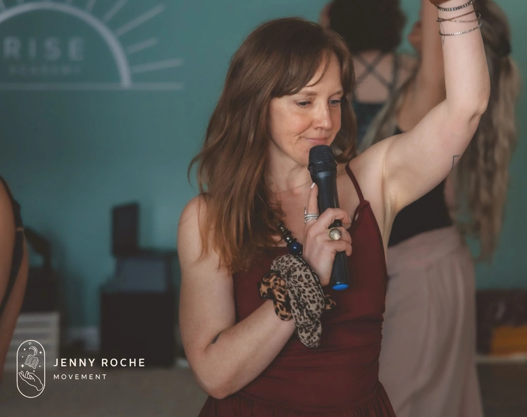 A woman with red hair holds a microphone close to her mouth with one hand and raises her arm in front of a teal wall. She wears a sleeveless dress with a leopard print patch near her waist and multiple bracelets on her wrist.