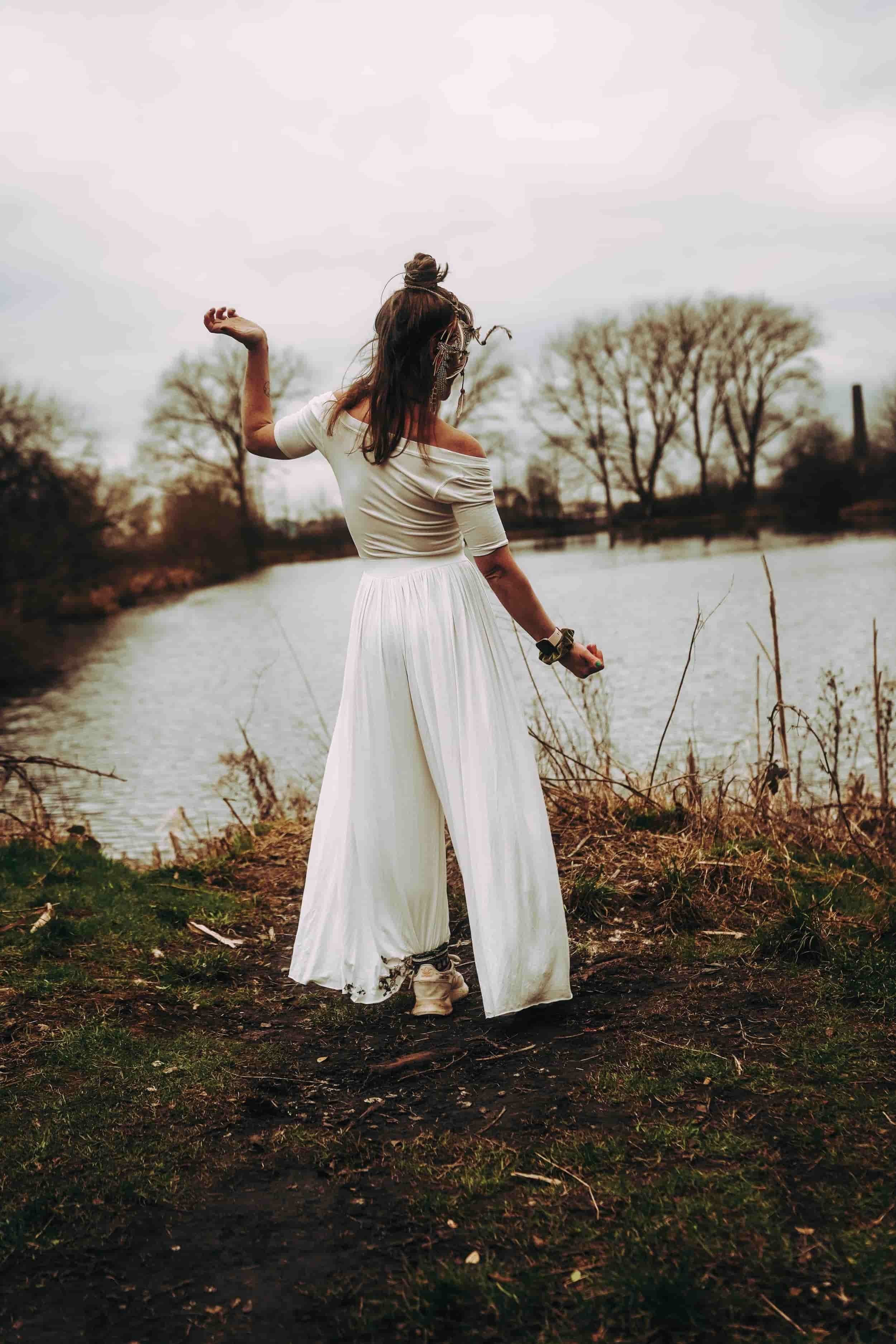 Jenny Roche, Dressed in long white flowing trousers stands in front of a lake, dancing and swaying