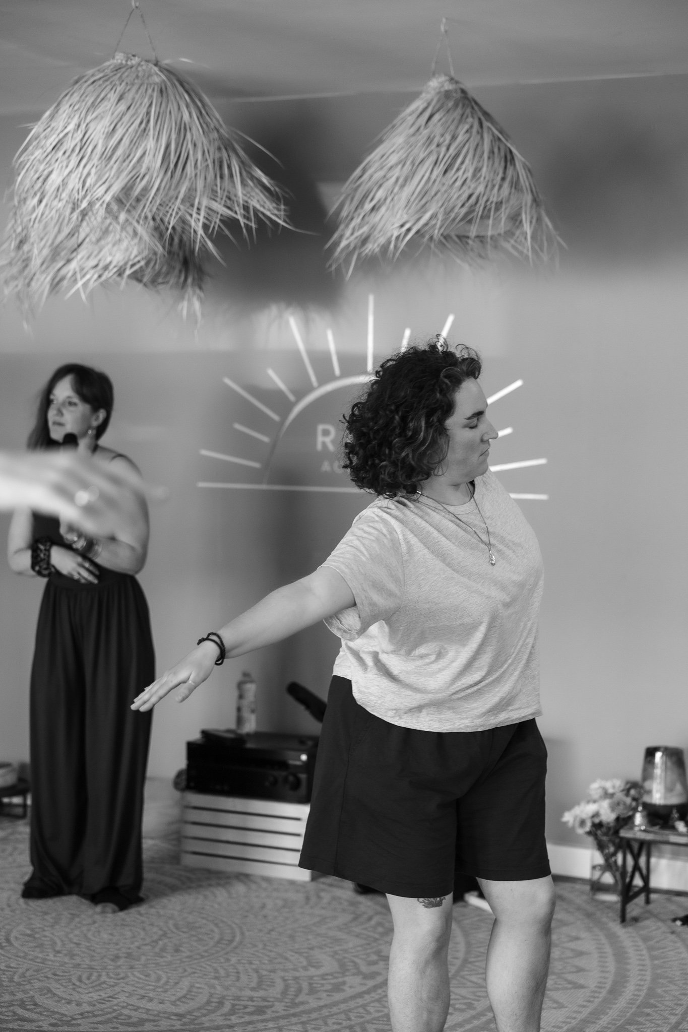 Two women standing in a room, one in the foreground with eyes closed and arms stretched out, the other in the background with arms crossed, both in a yoga or dance pose. The room has hanging ceiling decorations and a partial view of a floral table arrangement.