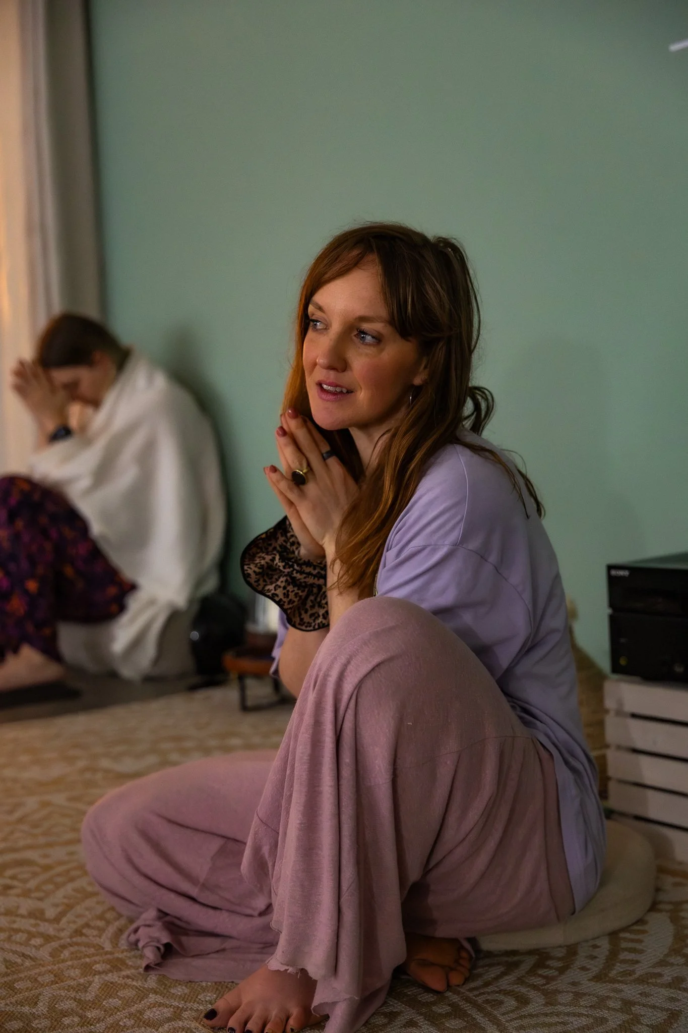 A woman with reddish-brown hair sitting on a cushion on the floor, clasping her hands near her face, with a contemplative expression. In the background, another person is sitting with their head bowed, wrapped in a blanket.