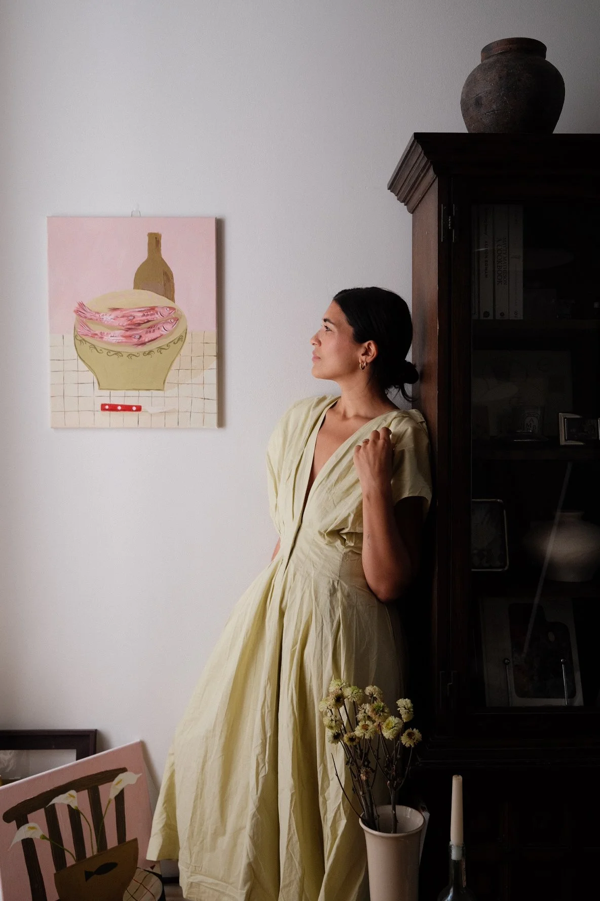 A woman wearing a light yellow dress leans against a wooden cabinet in a room with art and decorative items. There is a framed artwork of a jar with fish on the wall and a vase with flowers and a candle on a table.