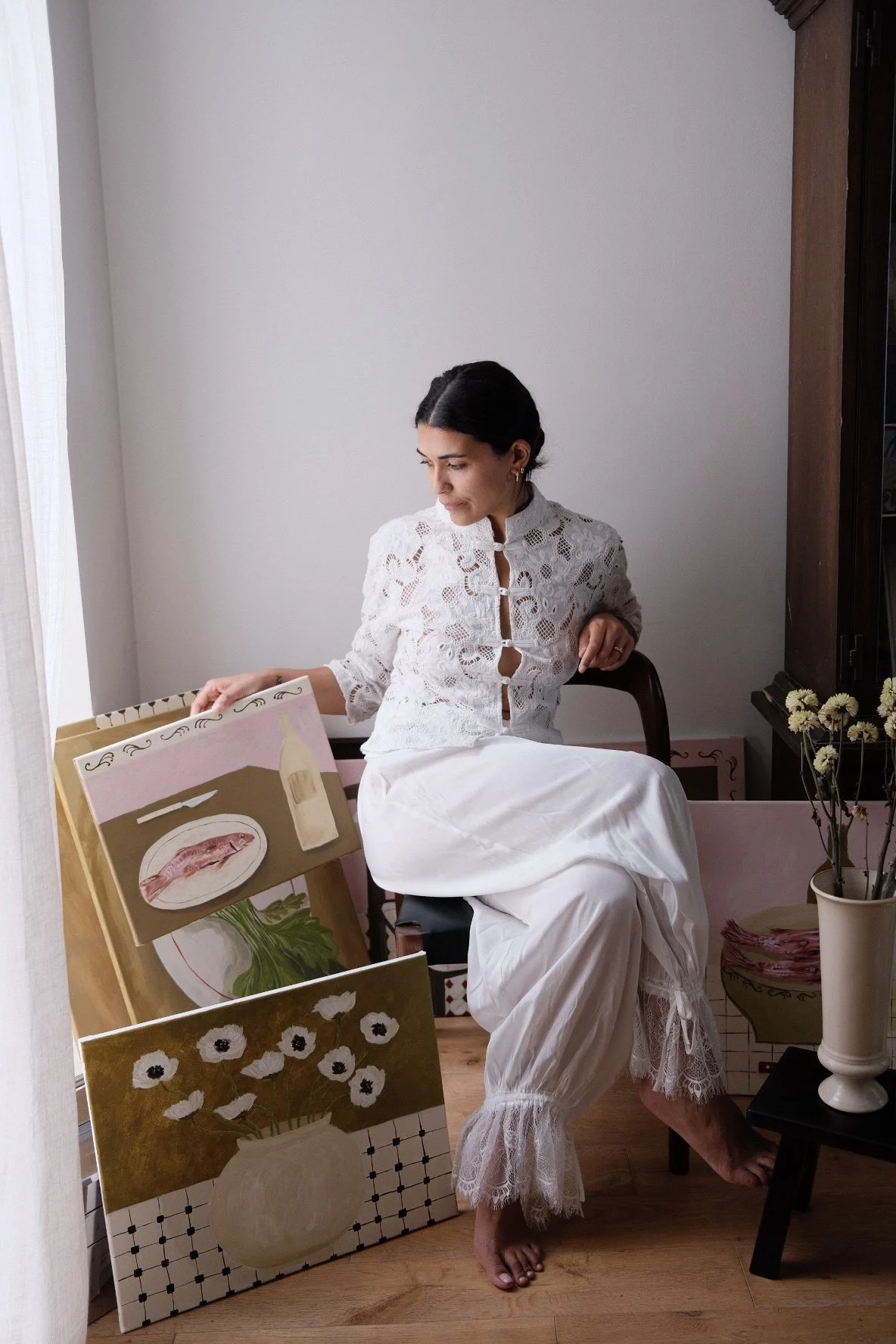 Woman sitting on a chair, wearing a white lace top and white pants, surrounded by paintings and flowers in a bright room.