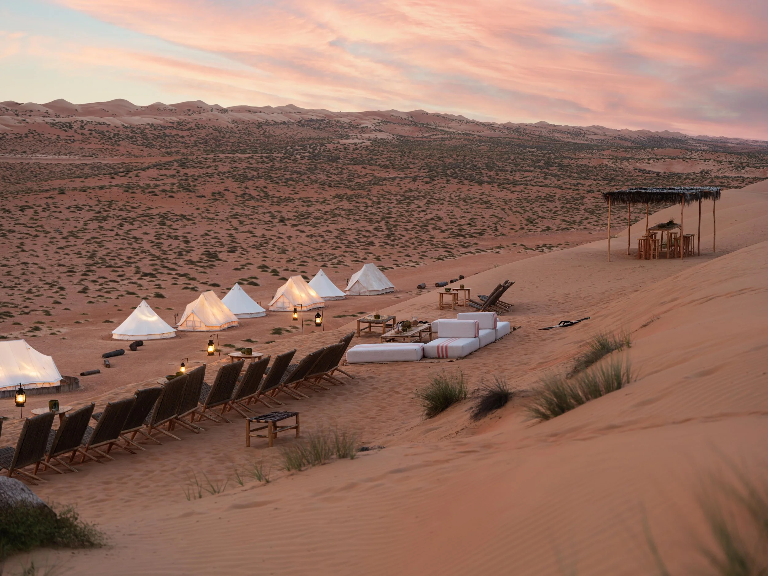 Desert camp with white tents, chairs, and tables set up on sand dunes under a sunset sky.