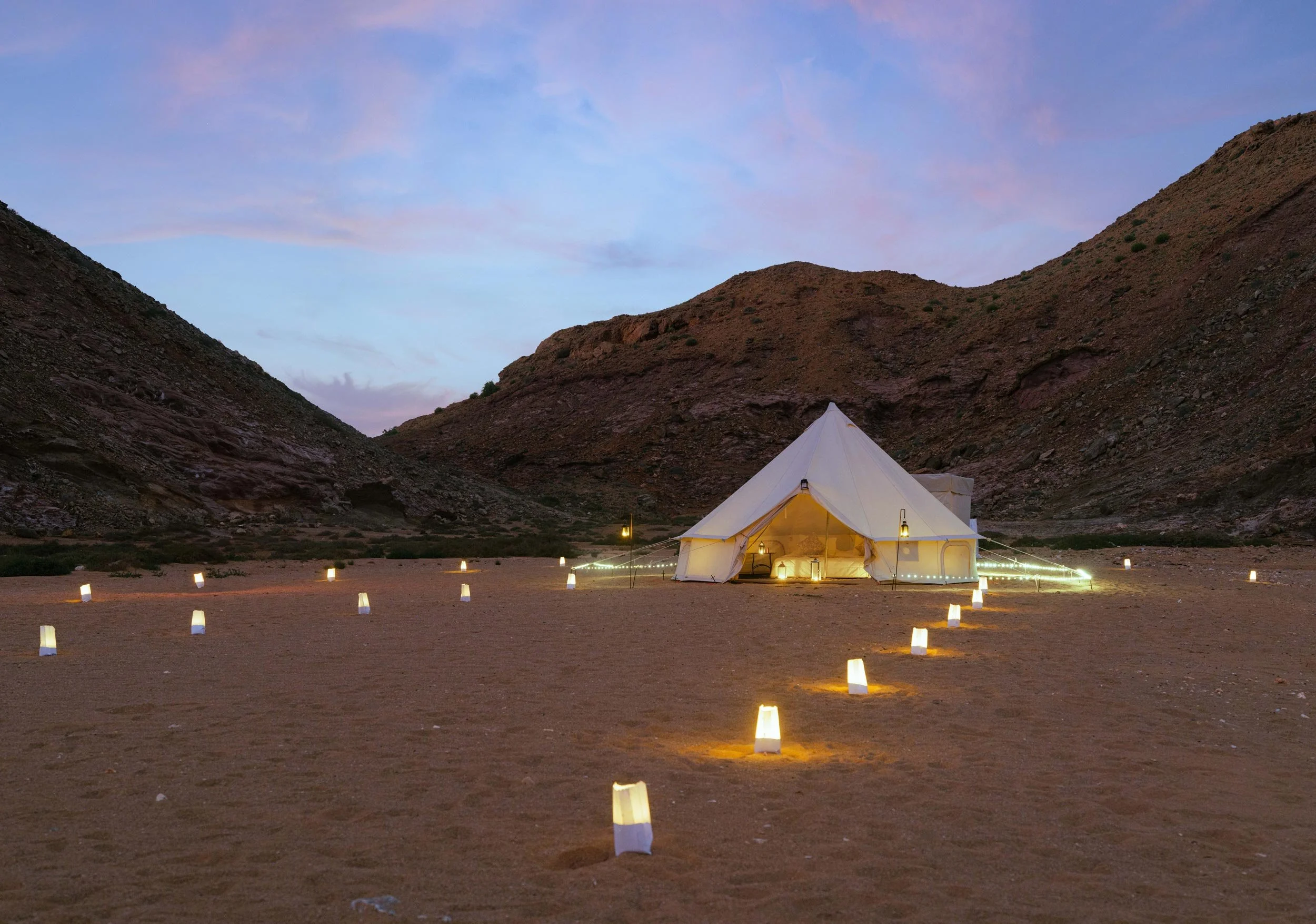 A large white tent set up in a desert valley surrounded by rocky hills, with string lights and lanterns illuminating the area at dusk in secluded beach of bandar al khiran - Oman.
